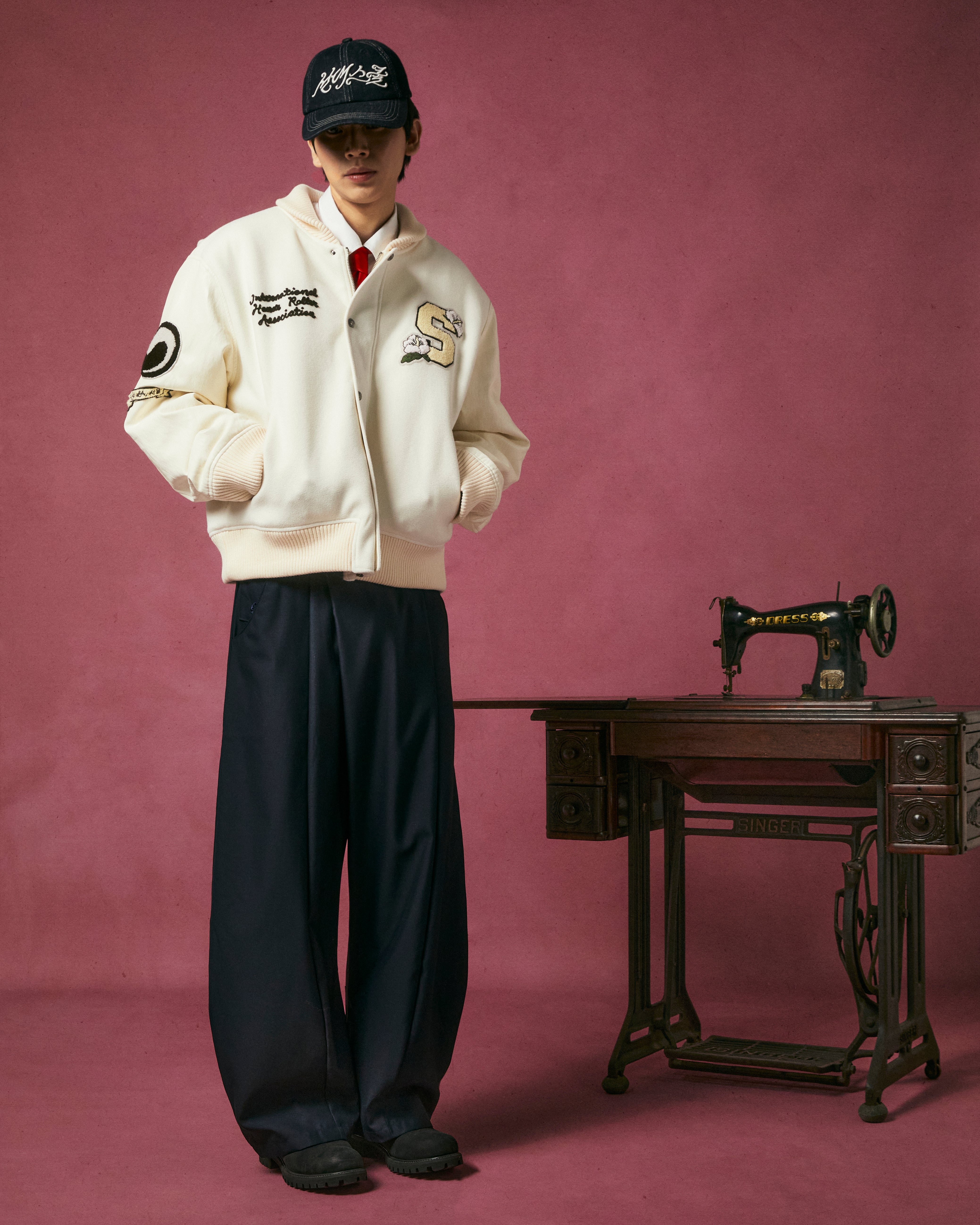 A person in Sundae School’s IVORY VARSITY JACKET with vegan leather sleeves, black wide-leg pants, and a black cap stands by a vintage sewing machine on a dark wood table against a pink backdrop—highlighting unisex streetwear style.