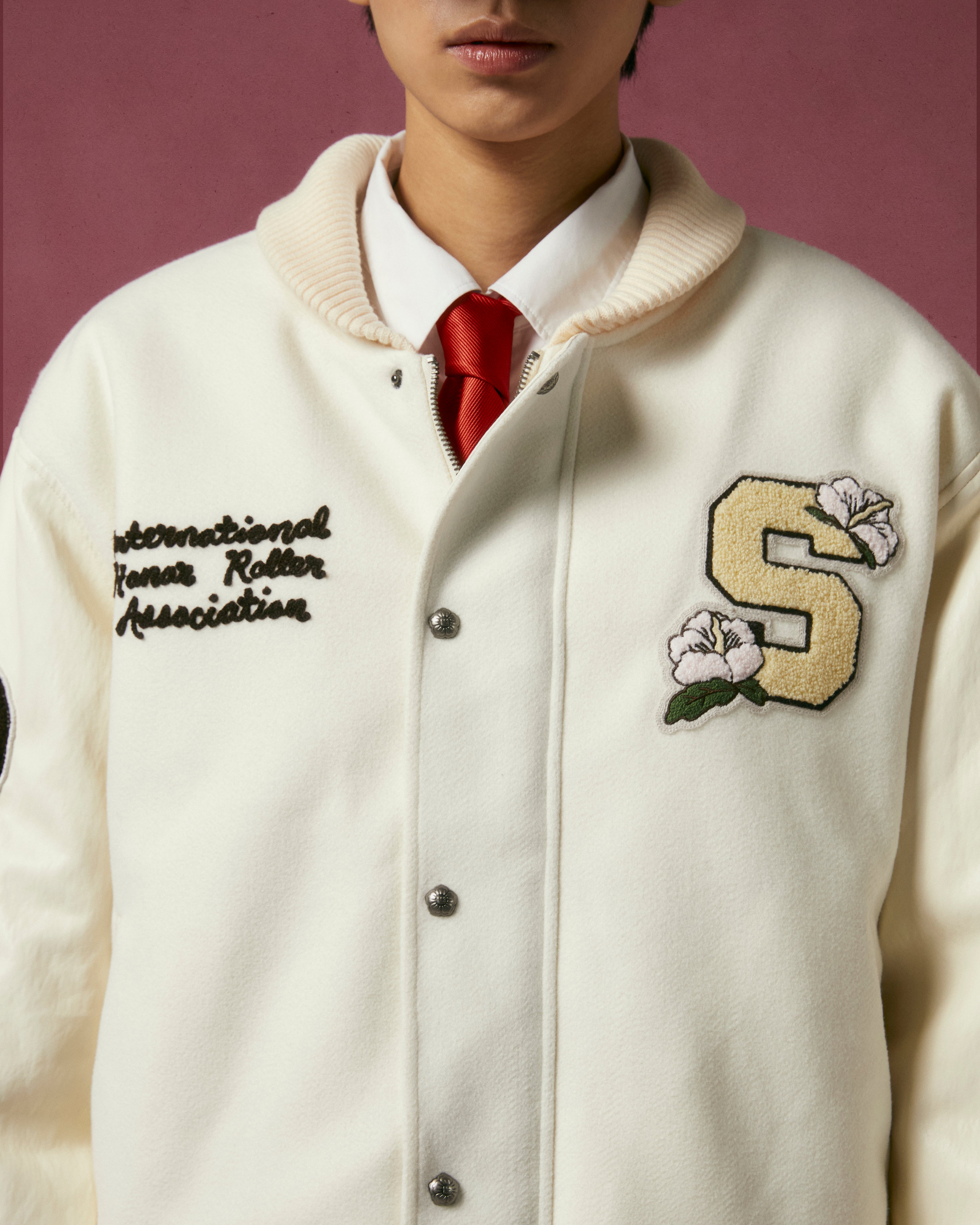 Someone wears the Sundae School IVORY VARSITY JACKET, with vegan leather sleeves, embroidered flowers, a big yellow S, and 