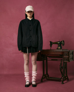 A person stands against a pink background wearing the Sundae School KNOT TIE TOWN JACKET with a pleated skirt, white leg warmers, and black shoes, next to a vintage Singer sewing machine on a wooden table.