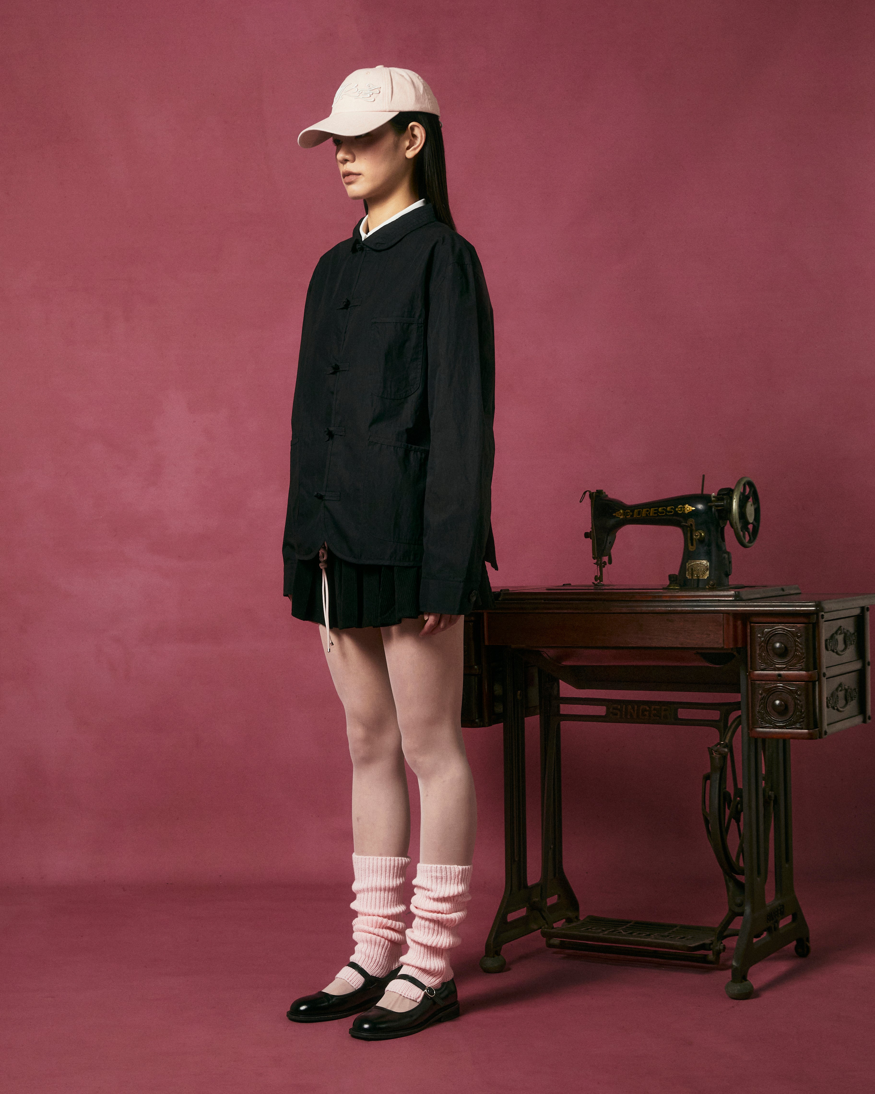 Wearing Sundae School's KNOT TIE TOWN JACKET, a pleated skirt, white cap, pink leg warmers, and black shoes, a model stands by an antique sewing machine against a pink background for a modern spring look.