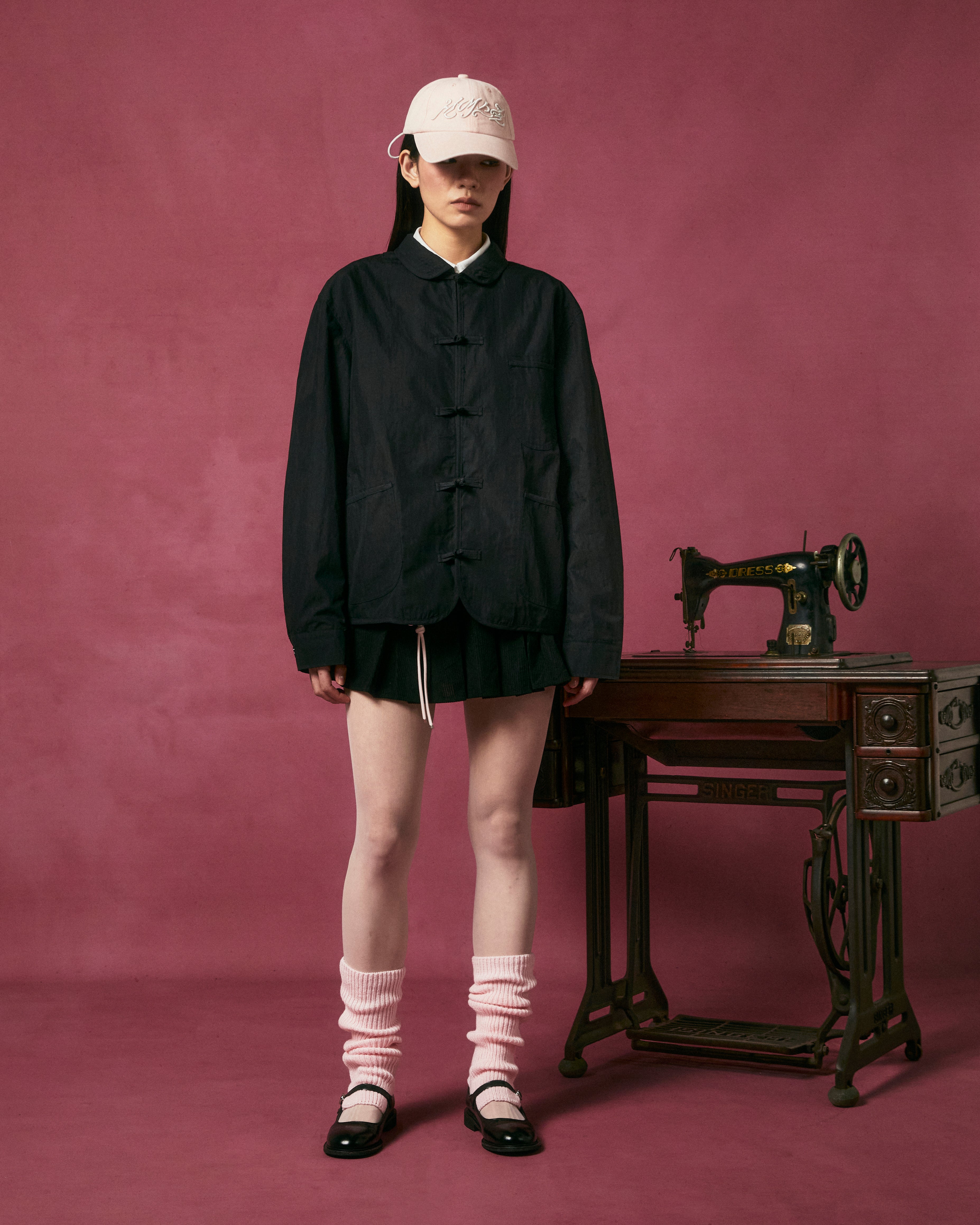 A person stands in front of a vintage Singer sewing machine, wearing Sundae School’s KNOT TIE TOWN JACKET, black shorts, a white cap, white socks, and black shoes against a dark pink background.