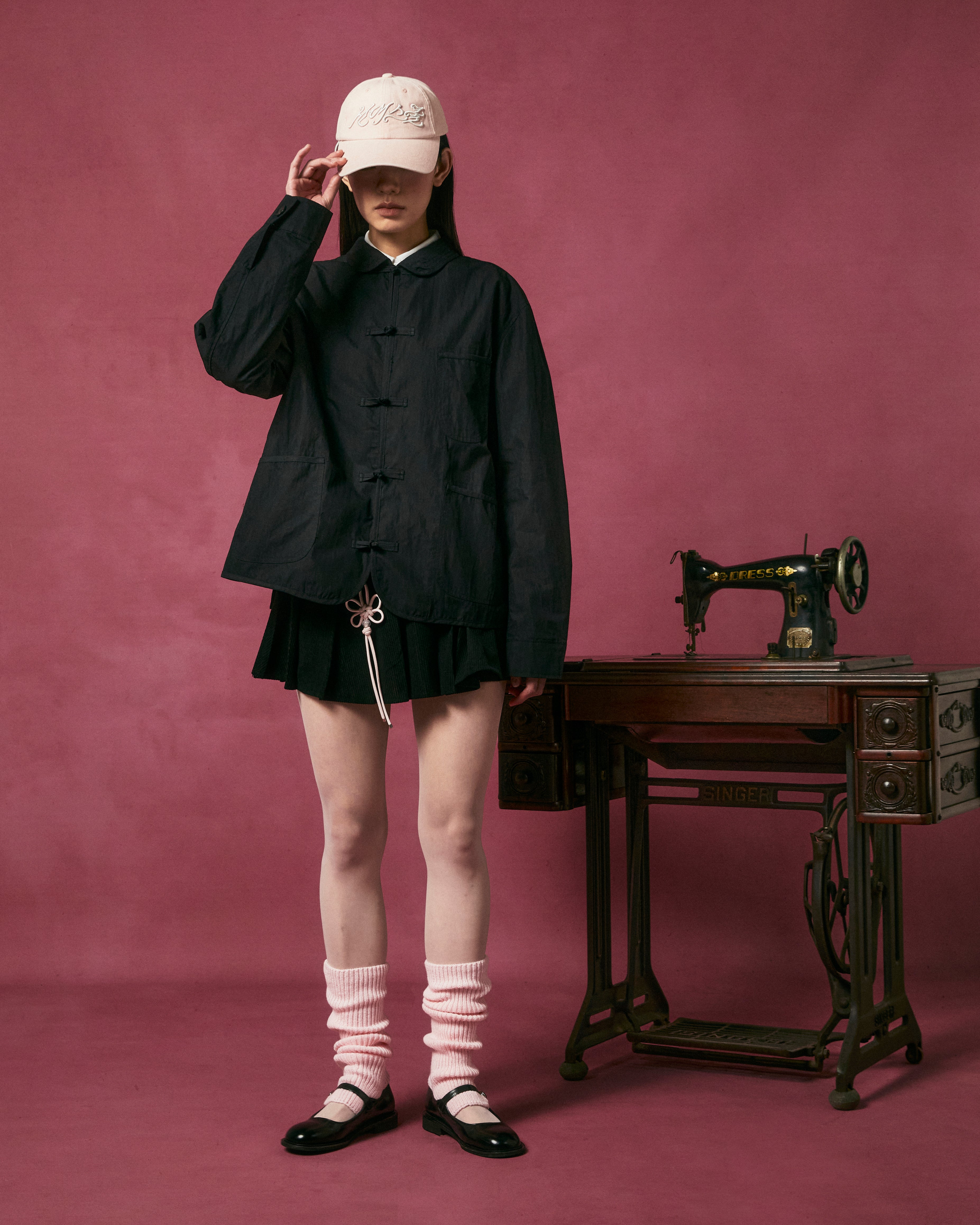 Wearing the KNOT TIE TOWN JACKET by Sundae School, a person in a black skirt, white hat, socks, and black shoes stands by a vintage sewing machine on a table with a pink background. Their face is partially hidden by the hat.