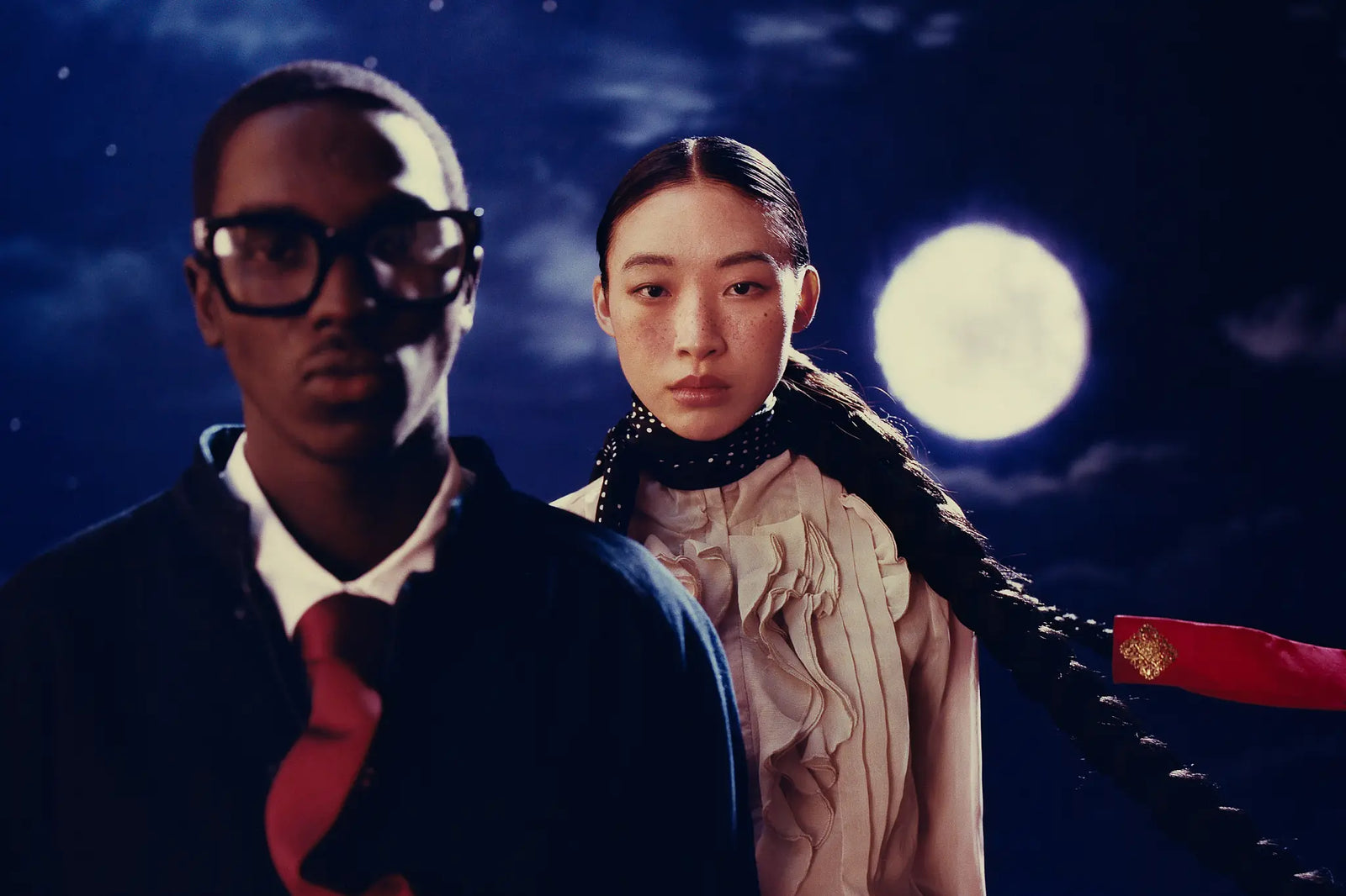 A man wearing thick black-rimmed glasses, a dark blazer, white shirt, and red tie stands beside a woman with long braided hair wearing a cream pleated top and a black collar, set against a night sky with a large full moon.