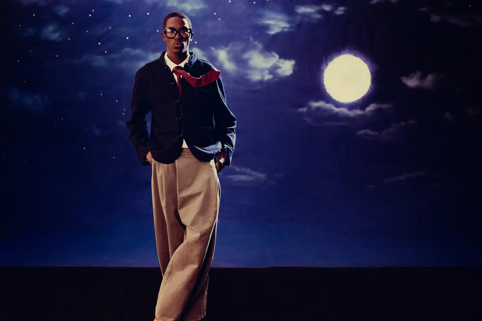 A man wearing a dark jacket, Sundae School Tan Hanbok Chino Pants, and a red neck scarf stands against a night sky backdrop with a large full moon and stars.