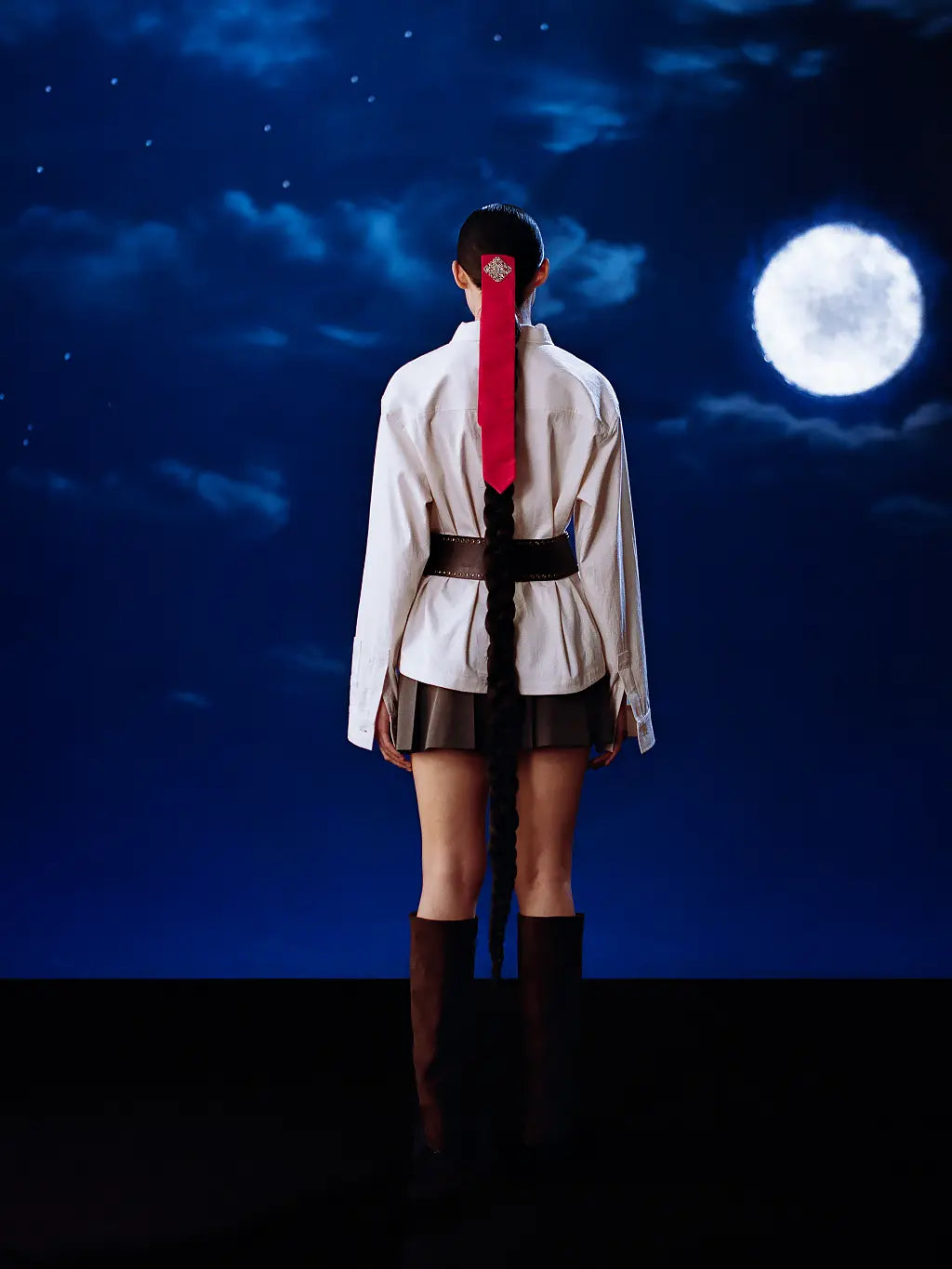 A model stands with their back to the camera wearing Sundae School Ivory China  Collar Knot  Shcaket, a dark pleated mini skirt, and dark brown knee-high boots. Their hair is styled in a long braid with a bright red ribbon accent.