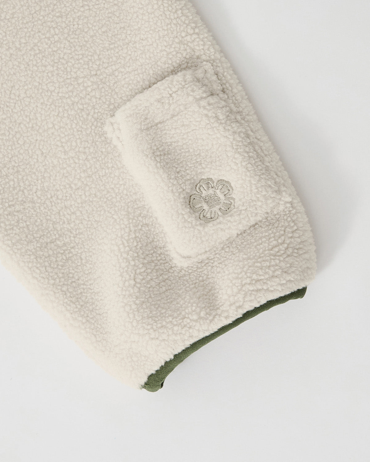 A close-up of Sundae School’s TAUPE TEARDROP KNOT FLEECE shows a cream-colored sleeve with a small rectangular pocket, an embroidered flower near the cuff, and an olive green inner edge.