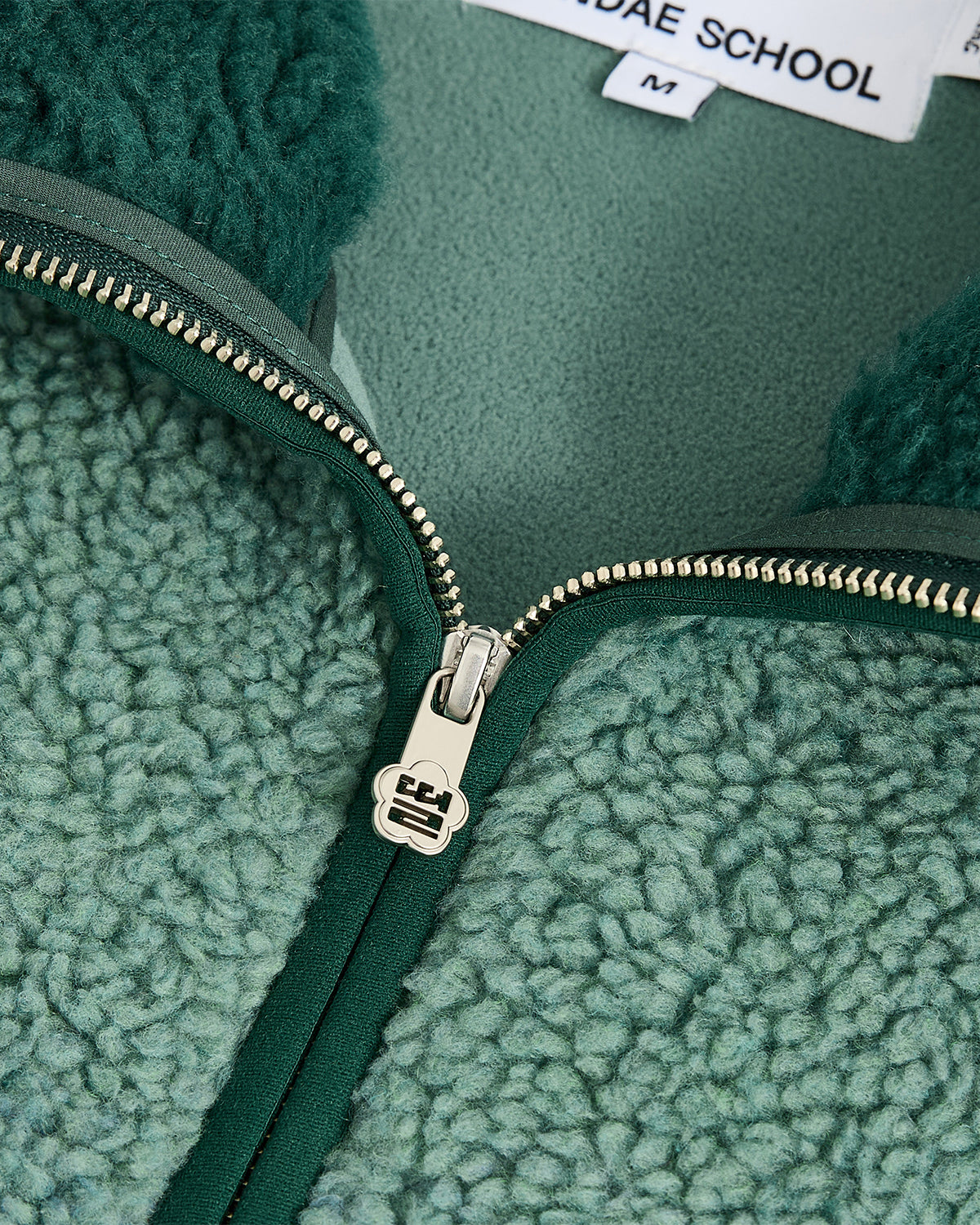 Close-up of a teal TEAL FIVE PEAKS FLEECE by Sundae School with a silver zipper partially unzipped, revealing soft, embossed pile fleece and a clothing label showing “Sundae School” and the size “M”.