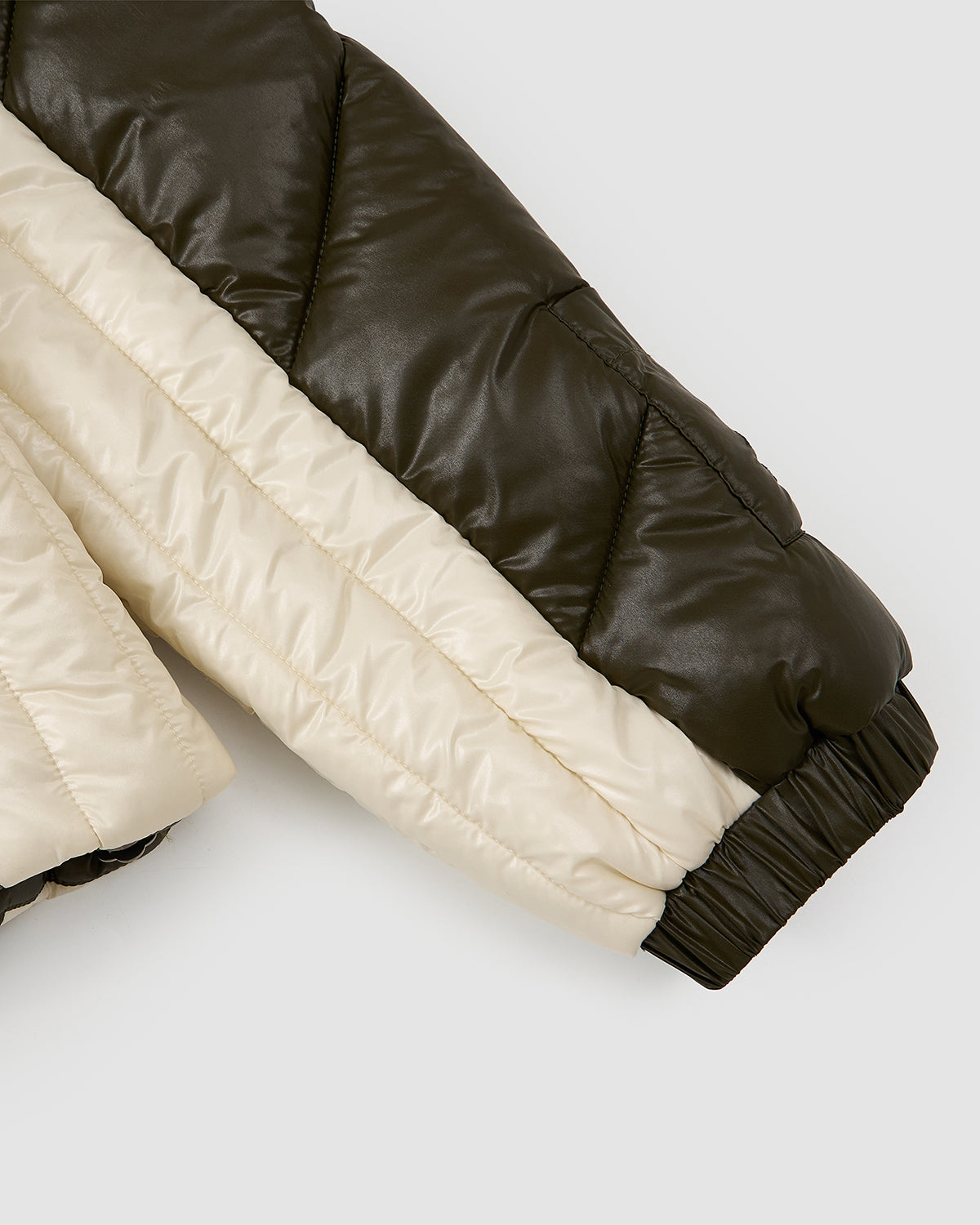 Close-up of the OLIVE OATMEAL MOCHI SPIRAL PUFFER by Sundae School, showing the puffy, lightweight sleeve in dark green and off-white color blocks with quilted stitching and an elastic cuff, laid on a light gray background.