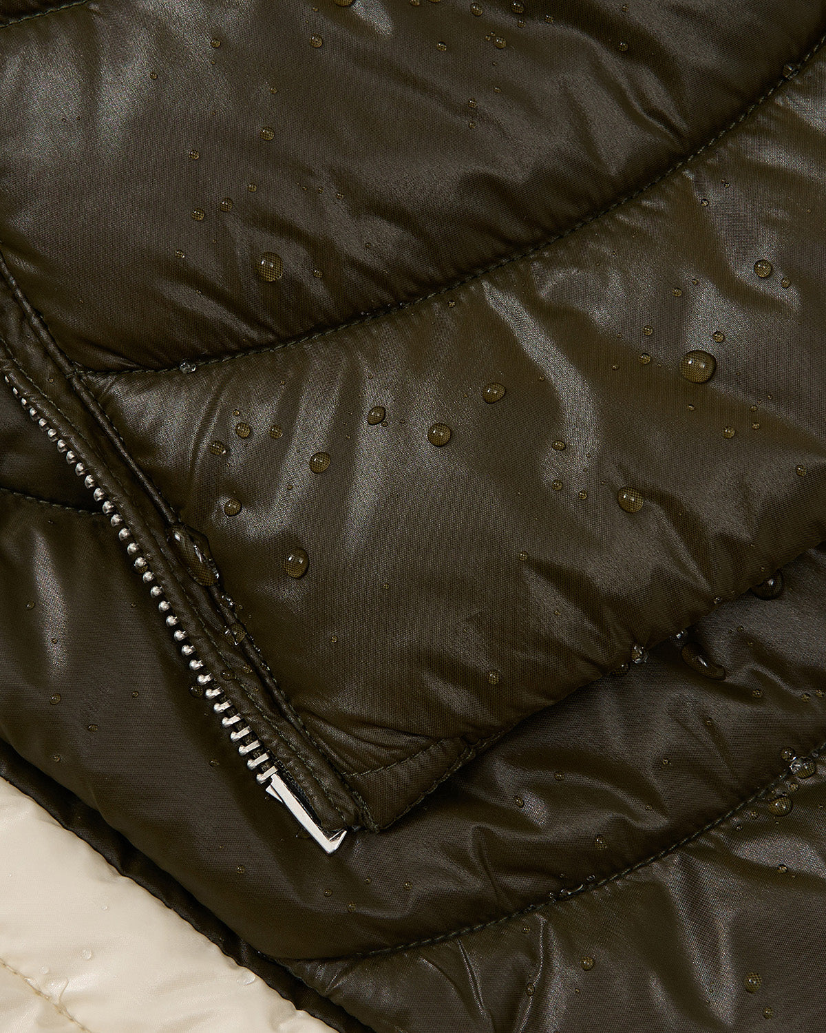 Close-up of the Sundae School OLIVE OATMEAL MOCHI SPIRAL PUFFER, featuring a shiny, quilted waterproof surface with water droplets and a silver zipper. A hint of light fabric appears in the corner.