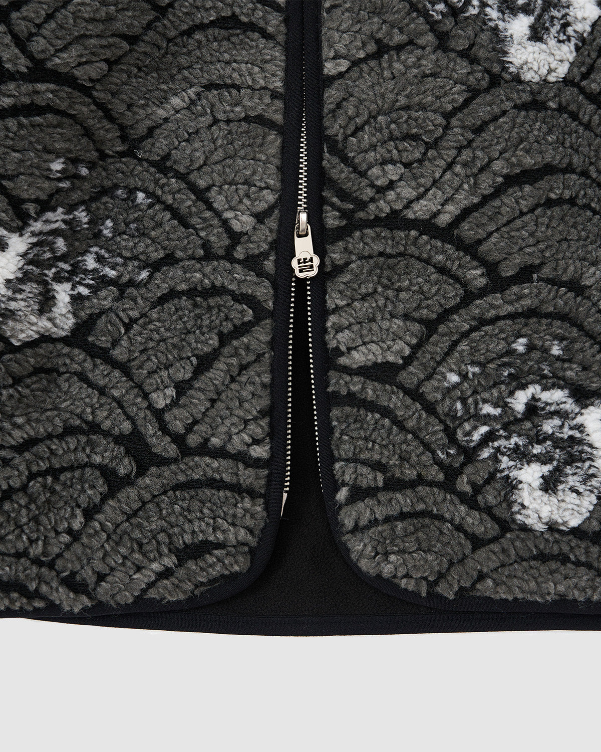 Close-up of the Sundae School BLACK WAVE FLEECE, featuring a textured, wave motif jacquard fabric in dark and light gray with a scalloped pattern and a silver zipper running vertically down the center.