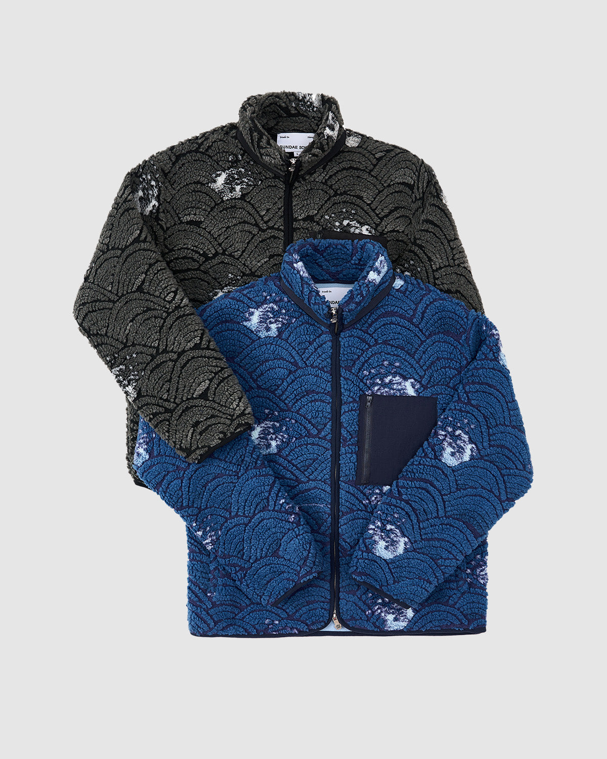 Two Sundae School BLACK WAVE FLEECE jackets—one in black/gray, one blue/navy—feature wave jacquard patterns and front zips. The blue version has a plain navy chest pocket. Both are shown layered against a light background.