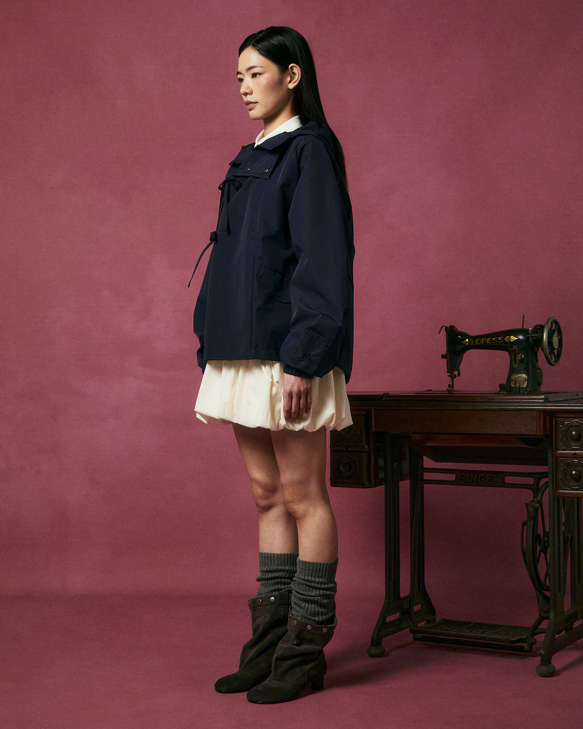 A woman stands in profile against a pink background, wearing the NAVY JEOGORI TIE SHELL JACKET by Sundae School, paired with a cream skirt, gray leg warmers, and brown boots beside an antique Singer sewing machine on a wooden table.