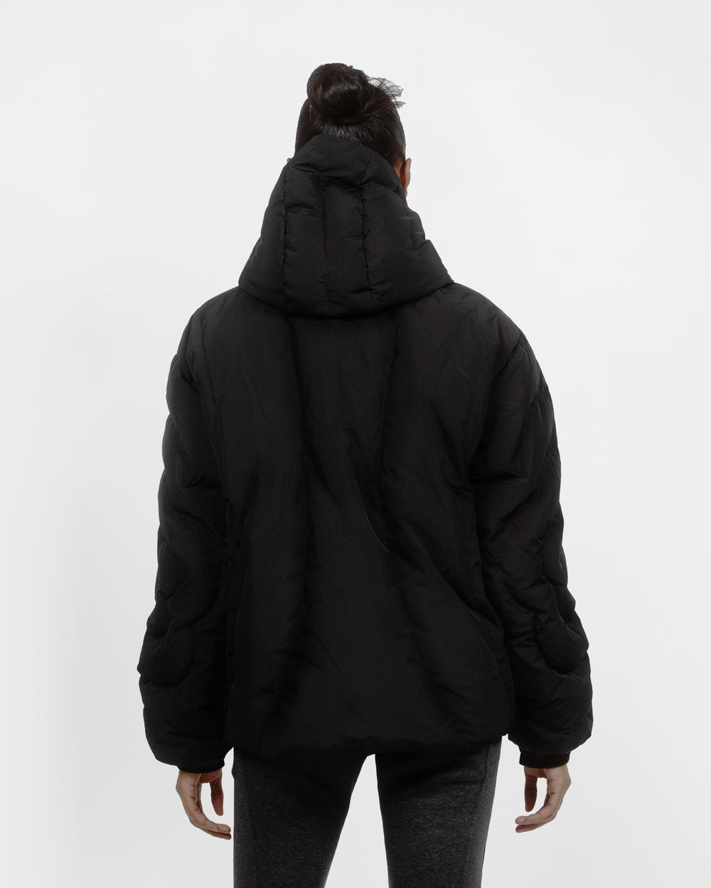 A person with dark hair in a bun models the Sundae School BLACK HEAVYWEIGHT MOCHI PUFFER, showcasing the back view and hood, paired with dark gray pants against a plain white background.