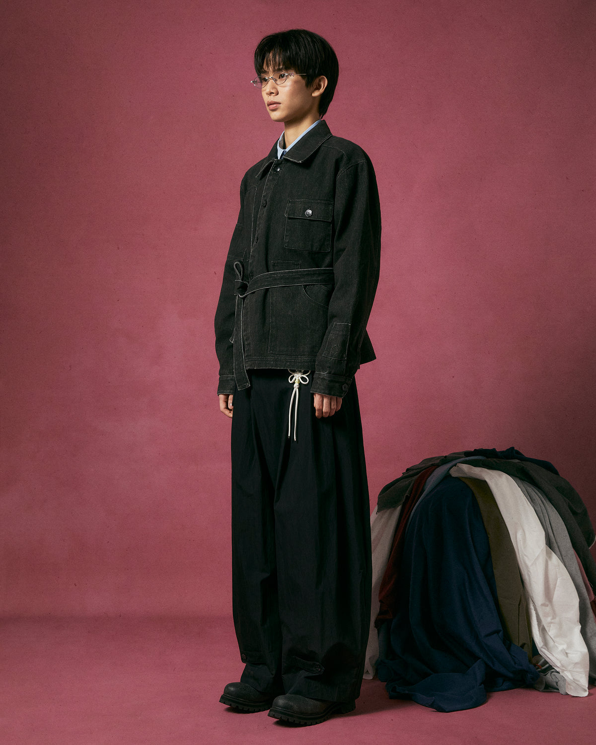 A person in glasses and a Sundae School BLACK ASYMMETRIC WORK JACKET with functional pockets and wide-leg pants stands on a pink background, with a pile of assorted clothing behind them to the right.