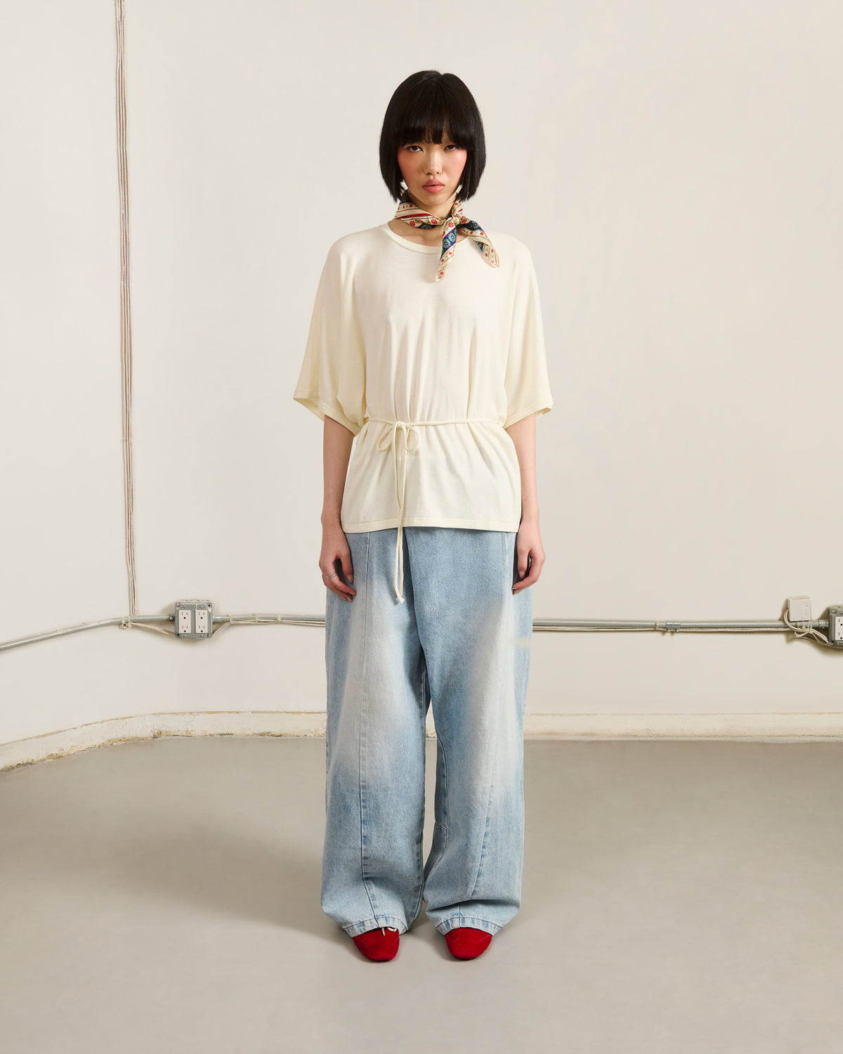 A person with short dark hair wears Sundae School SKY BLUE HANBOK DENIM PANTS, a cream belted top, red shoes, and a patterned scarf around their neck, standing against a plain wall with exposed electrical outlets and wires.