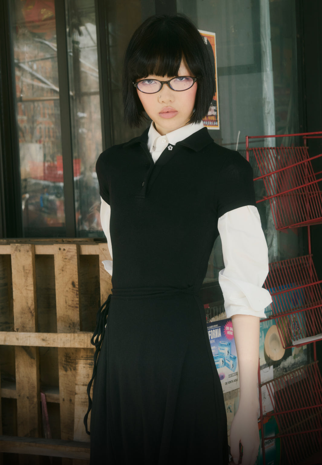 A vertical portrait of a model with a black bob and glasses wearing a layered white button-down and black short-sleeved polo, standing in an urban setting in front of a wooden pallet and a red wire rack.