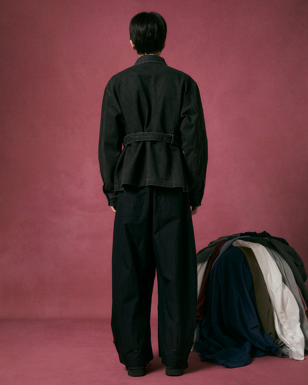 A person with short dark hair faces away, wearing Sundae School's BLACK ASYMMETRIC WORK JACKET and loose black pants against a pink background. To the right is a pile of assorted folded clothes in various colors.