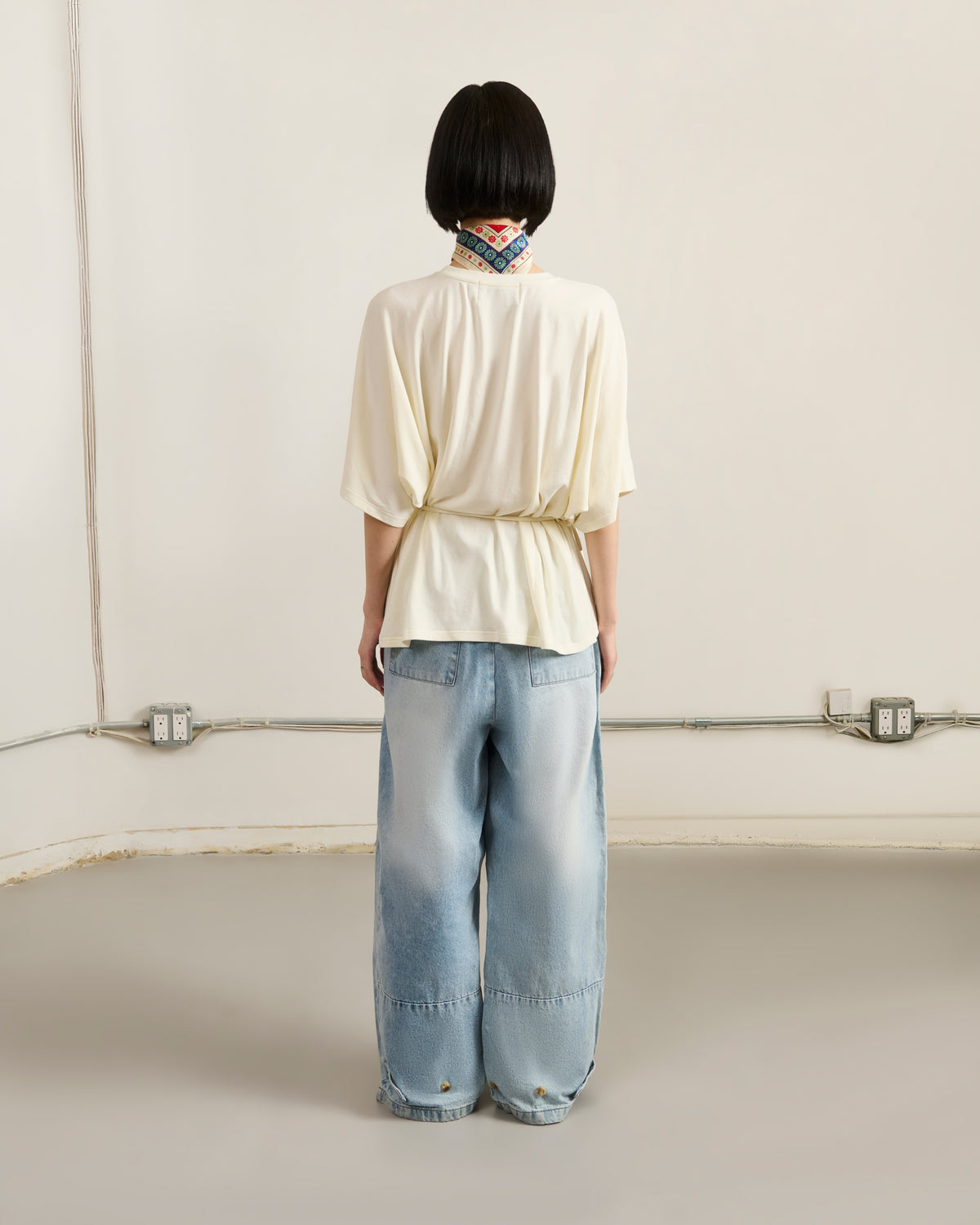Facing a plain wall with exposed outlets and wires, a person with short black hair stands with hands by their sides, wearing a cream top and Sundae School's SKY BLUE HANBOK DENIM PANTS featuring wide balloon legs.