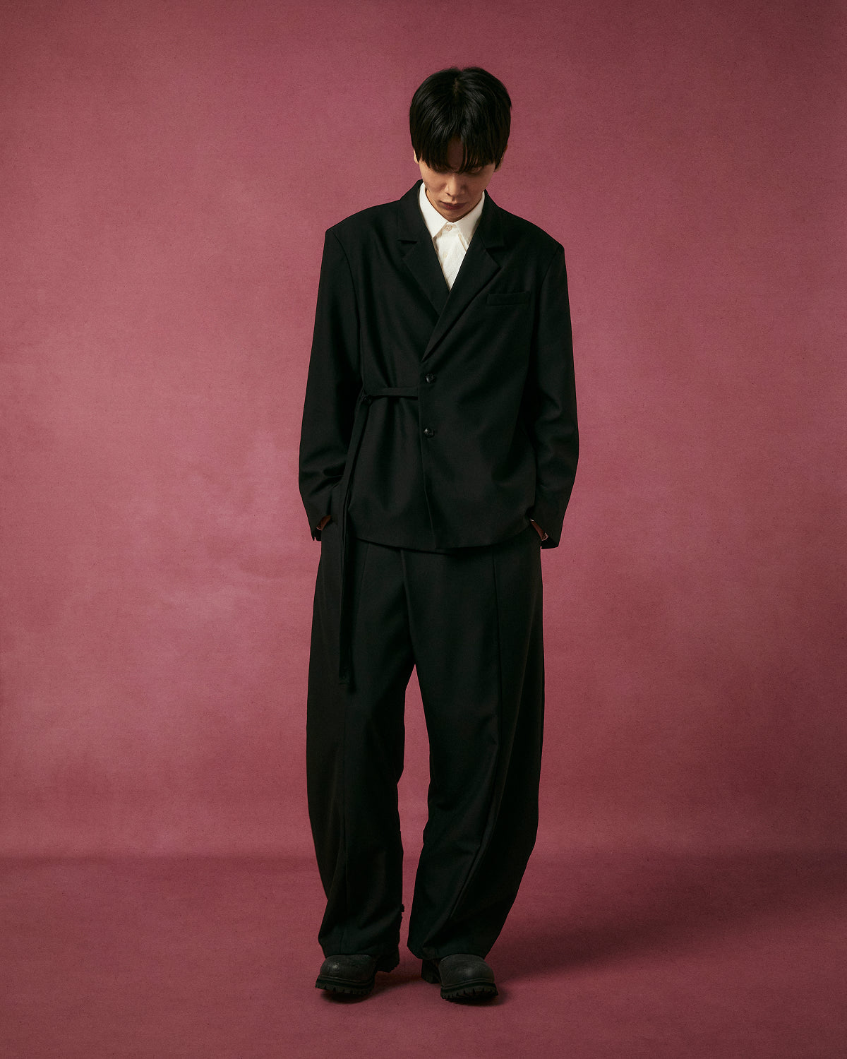 A person stands on a burgundy backdrop wearing Sundae School’s BLACK PLEATED HANBOK SLACKS as part of a black suit with a white shirt and black shoes, looking down with hands in pockets.