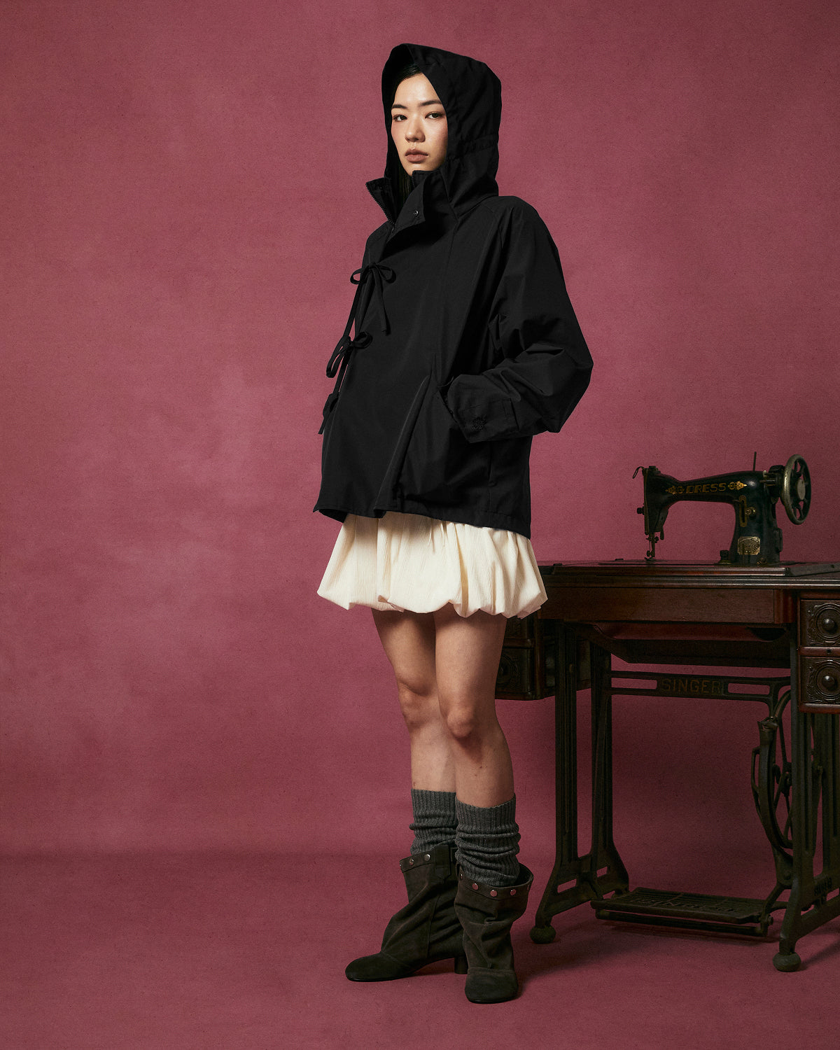 A person wearing the Sundae School BLACK JEOGORI TIE SHELL JACKET—a black, water-resistant hooded outerwear piece inspired by the Korean jeogori—paired with a white skirt, gray leg warmers, and brown boots stands next to an antique sewing machine against a pink background.