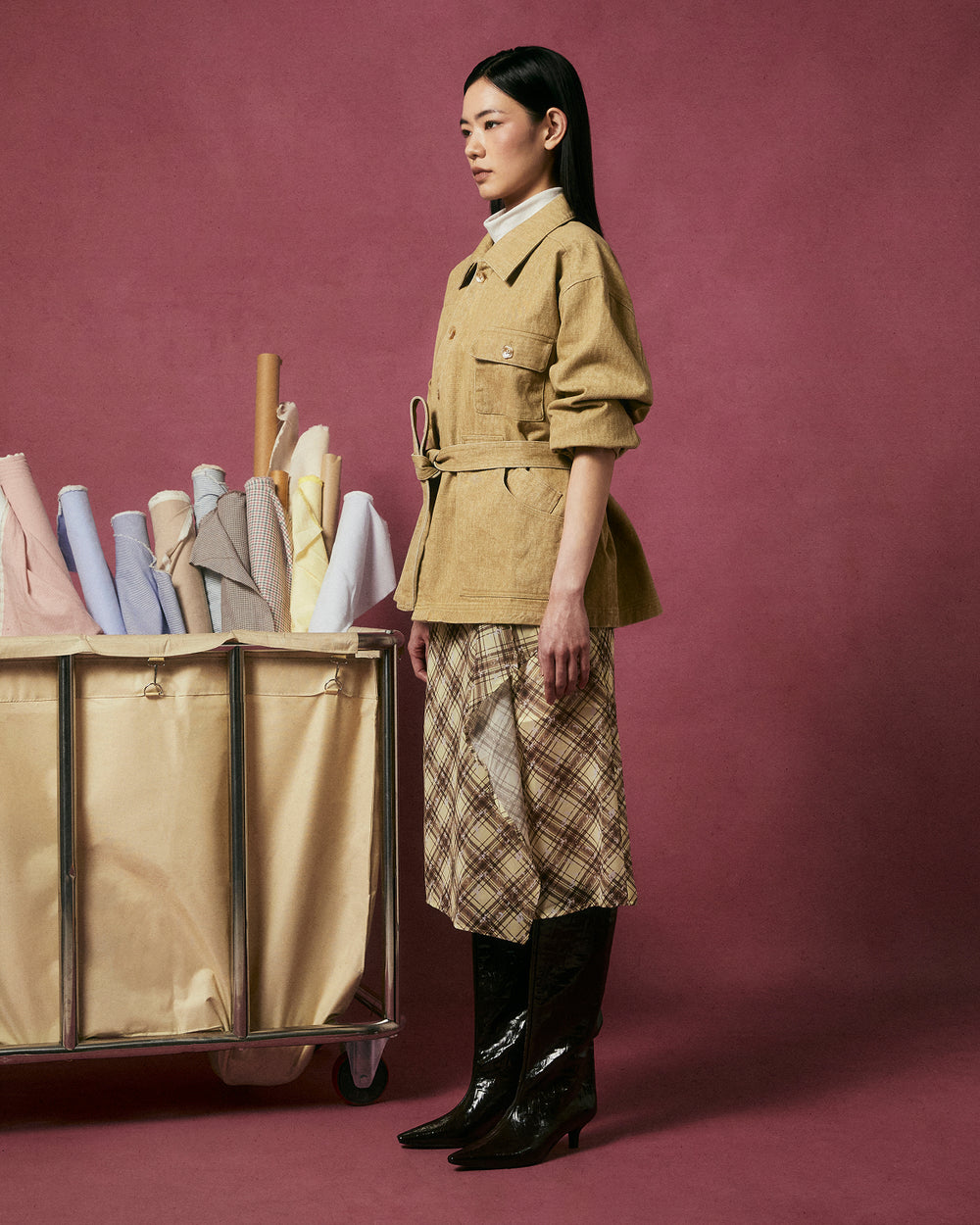 A woman in profile stands against a pink background, wearing Sundae School's TAN ASYMMETRIC WORK JACKET, a plaid skirt, and black boots. Beside her is a rolling cart with colorful fabric rolls.