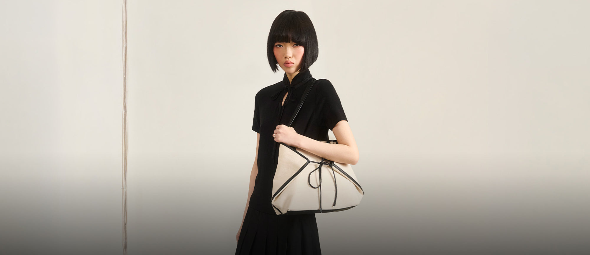 A horizontal web banner featuring a model with a black bob wearing an all-black ensemble and carrying a large cream-colored tote bag with black trim and a drawstring bow detail, set against a neutral off-white background.