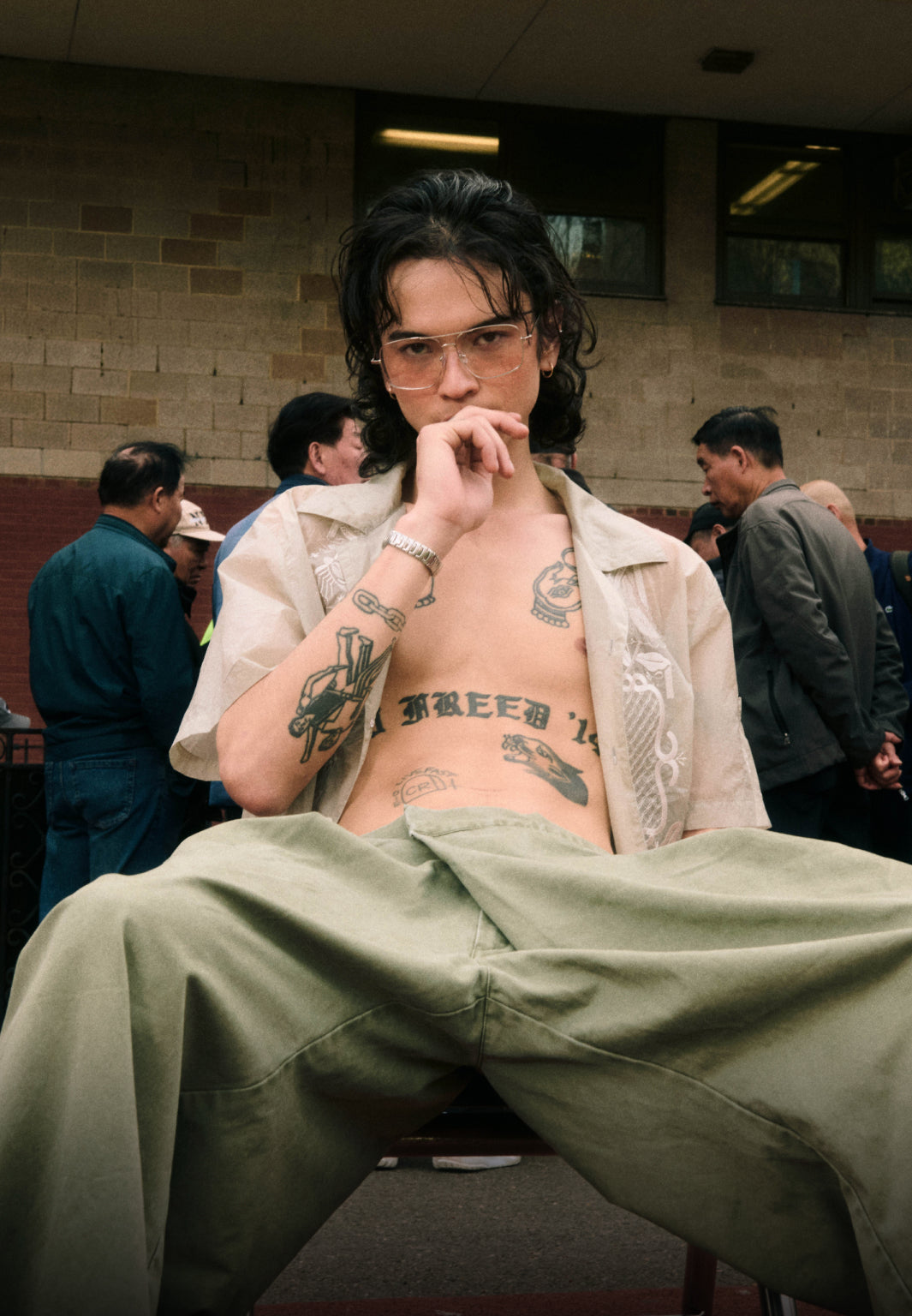 A vertical portrait of a model with long dark hair, glasses, and torso tattoos, seated with their legs spread wide. They are wearing an open, sheer embroidered cream shirt and baggy olive trousers, set against an outdoor urban background with bystanders standing near a brick building.