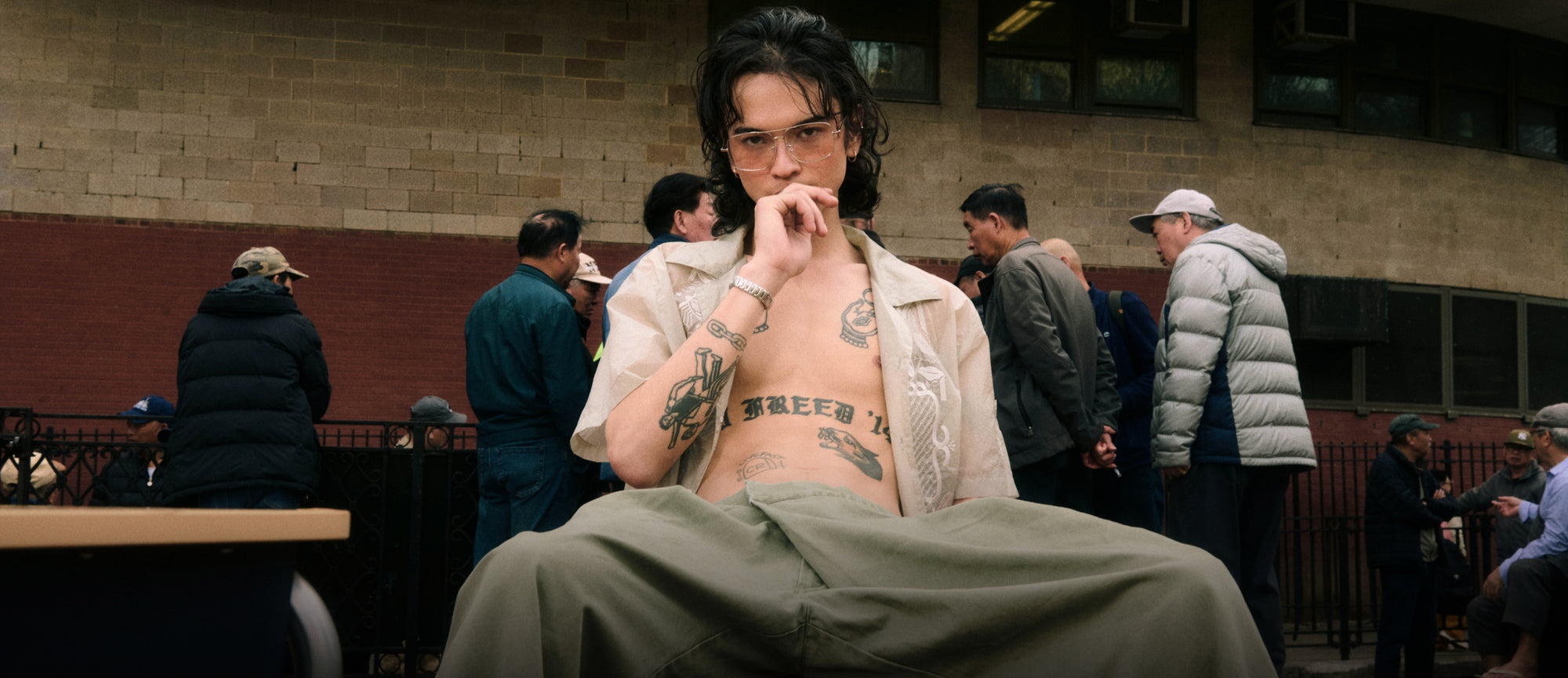 A horizontal web banner featuring a wide shot of a model seated outdoors in an unbuttoned, sheer embroidered cream shirt and wide-leg olive trousers. The low-angle composition highlights their tattoos and relaxed pose, with a crowd of people and a brick building filling the background space.