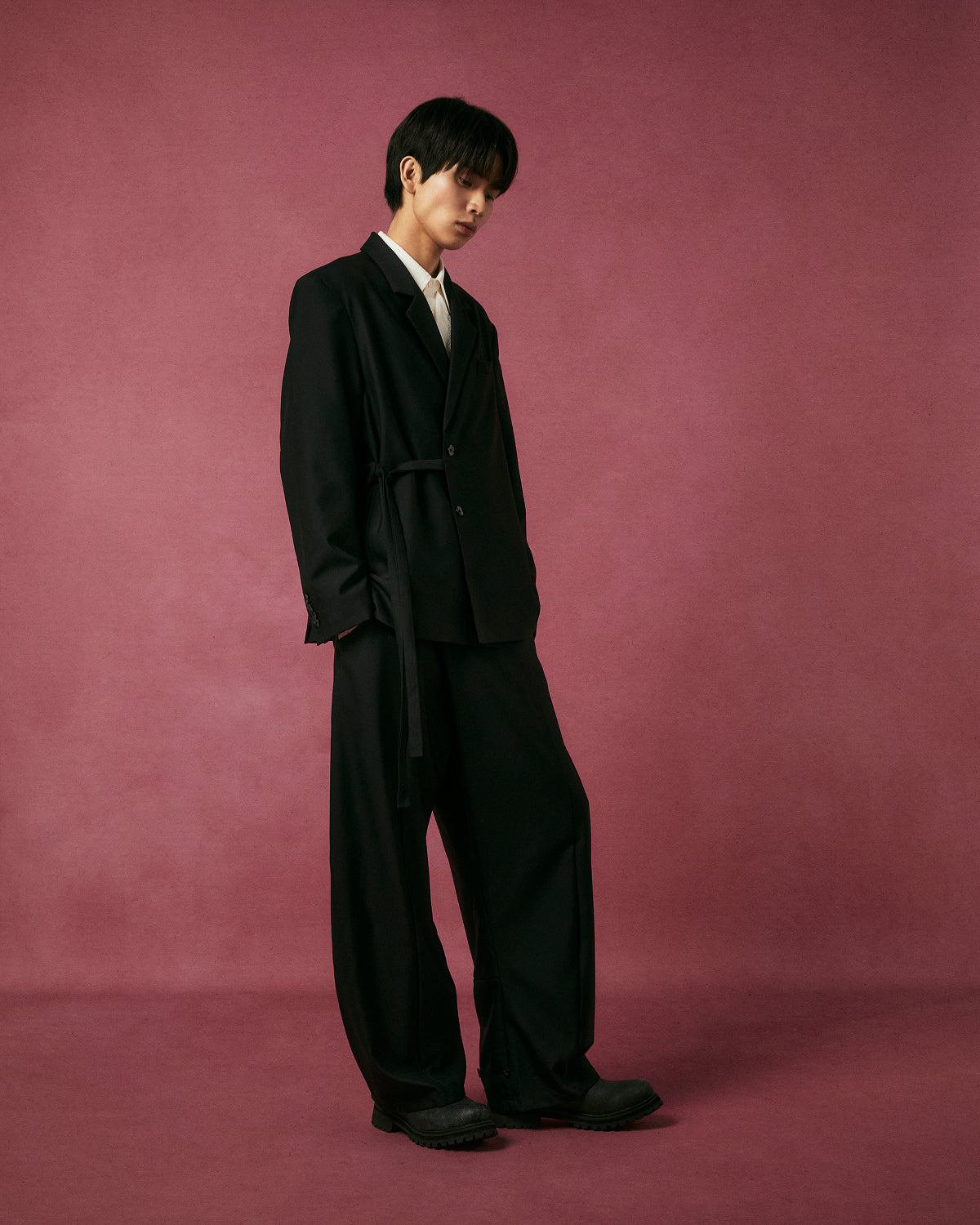 A person with short dark hair stands against a maroon background, wearing Sundae School's BLACK PLEATED HANBOK SLACKS, a white collared shirt, black shoes, and an oversized black suit. They pose in a relaxed, slightly tilted stance.