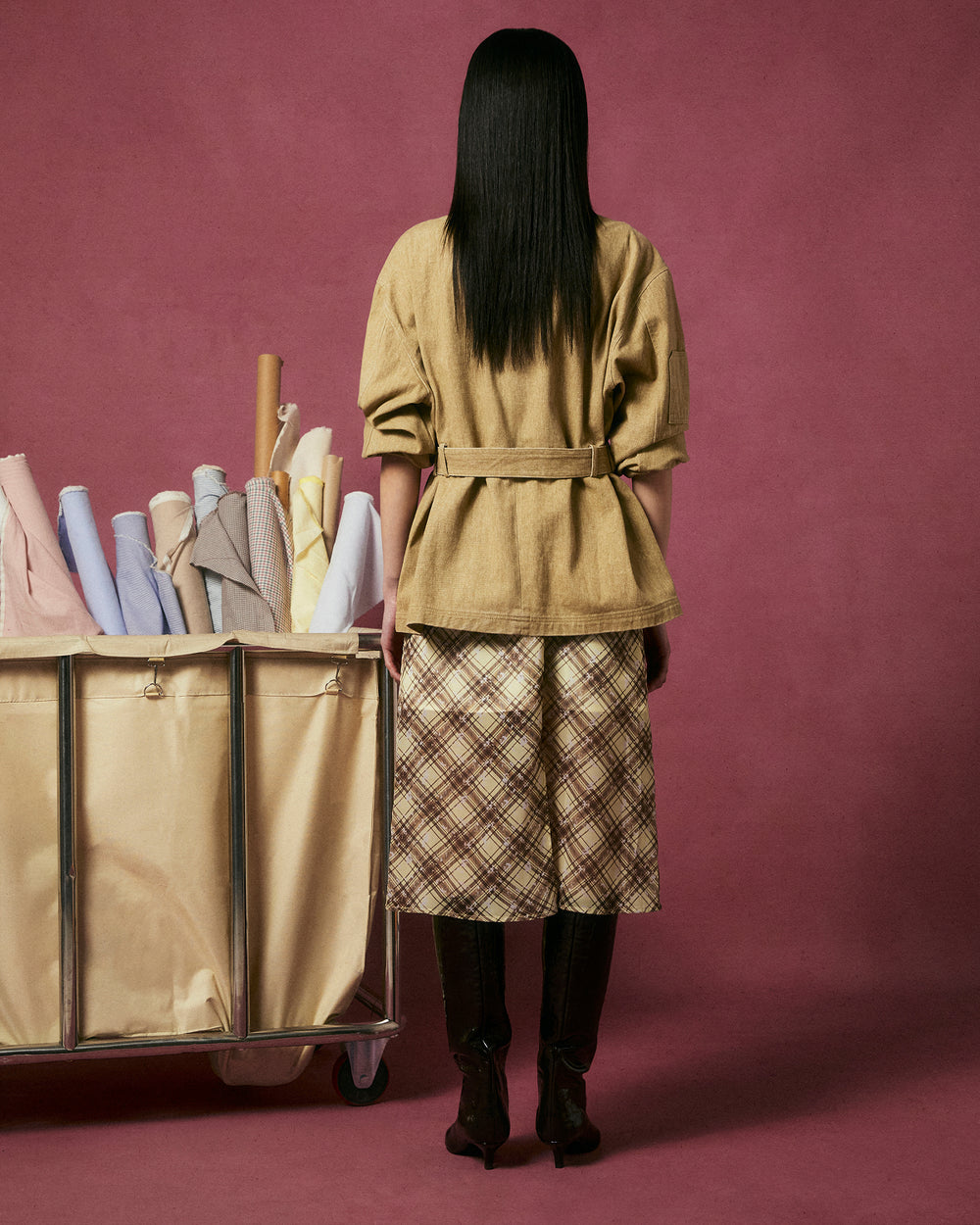 A person with straight, dark hair stands facing away, wearing the TAN ASYMMETRIC WORK JACKET by Sundae School and a plaid skirt near a beige fabric-filled cart, all set against a solid mauve background.