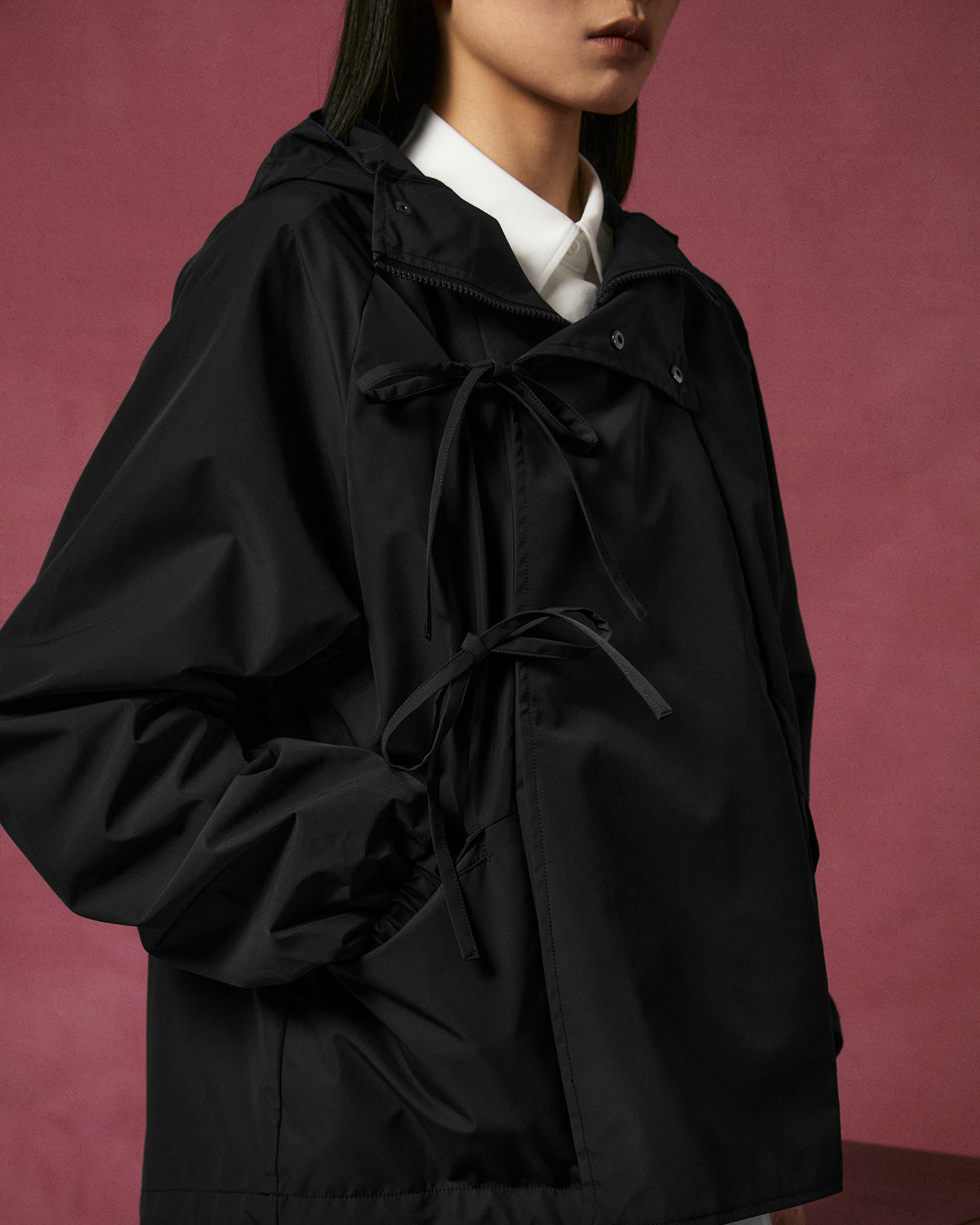 A woman models the BLACK JEOGORI TIE SHELL JACKET by Sundae School, a loose-fitting, water-resistant black hooded outerwear piece featuring tied bows, layered over a white collared shirt. Her lower face and upper body are shown against a muted pink background.