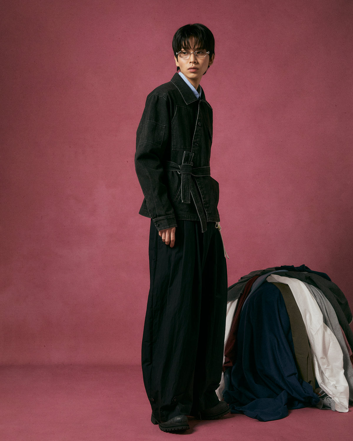 A person with short dark hair and glasses stands against a pink backdrop, wearing Sundae School's BLACK ASYMMETRIC WORK JACKET paired with wide black pants. A pile of assorted jackets lies on the floor beside them.