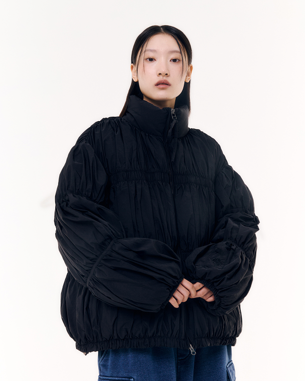 The BLACK PLEATED DUVET PUFFER by Sundae School is a black, long-sleeve jacket with pleats, a high collar, zipper closure, ruched texture, elastic cuffs, and a relaxed fit for cozy warmth.