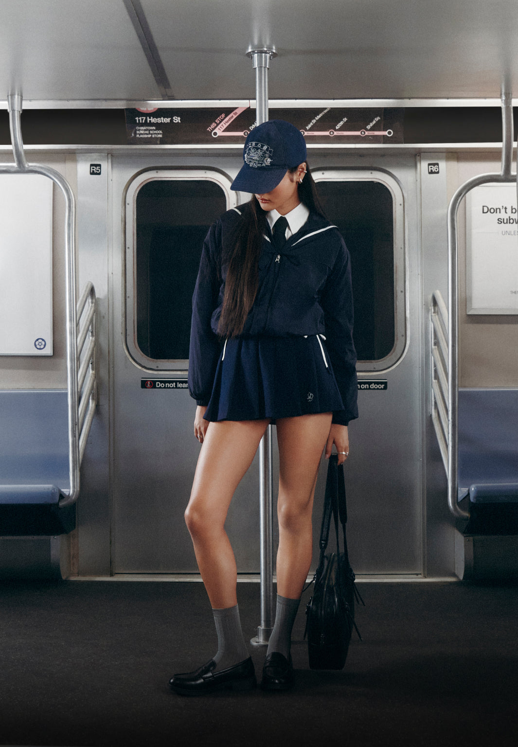 A vertical full-body shot of a model standing inside a subway car, holding onto a center metal pole. She is wearing a navy streetwear school-uniform look: a sailor-collared zip jacket over a white shirt and tie, a pleated mini skirt, gray socks, black loafers, and a navy baseball cap. The background features subway doors and an overhead transit map marked "117 Hester St."