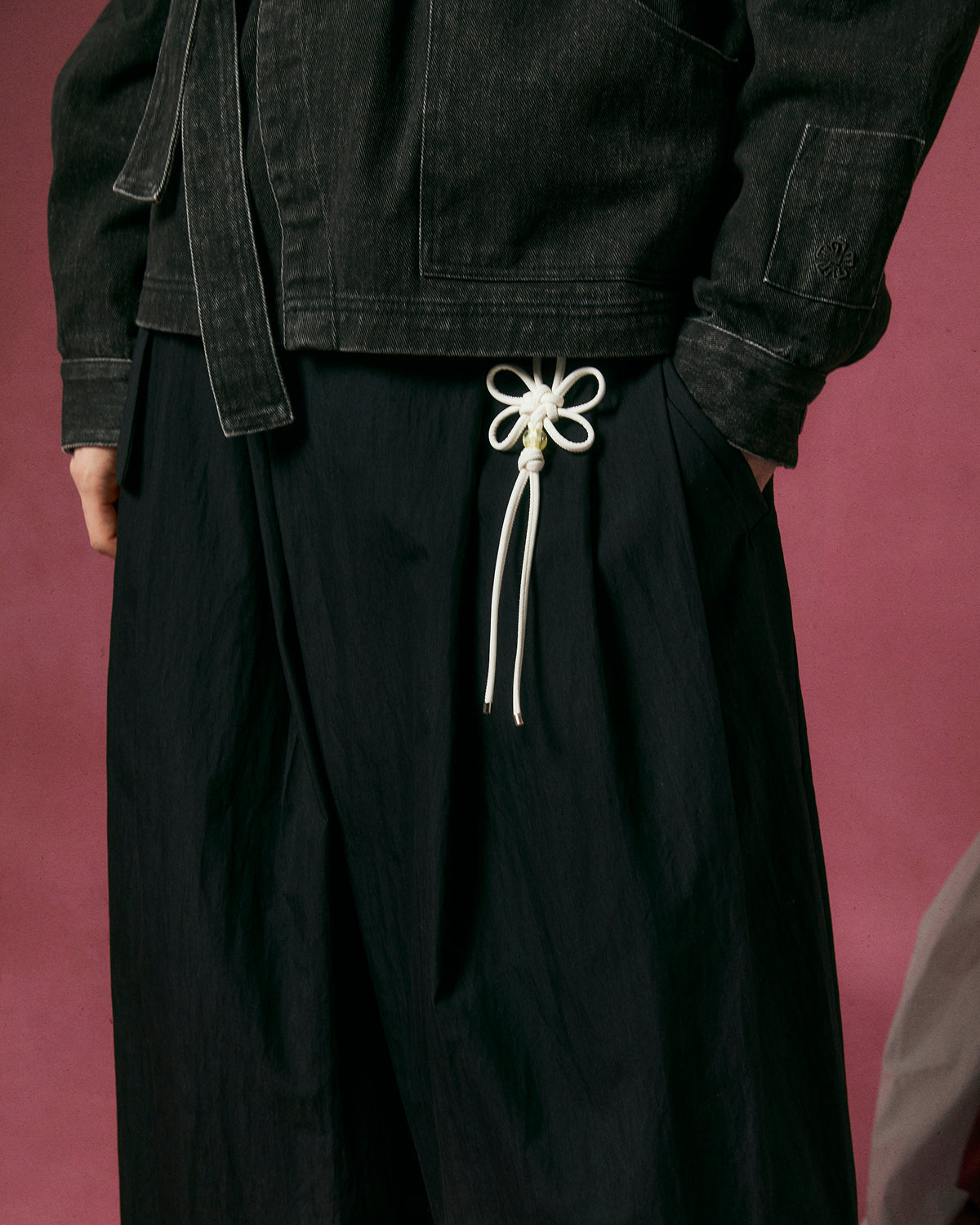 A person wears Sundae School's BLACK PLEATED PALAZZO PANTS with a matching jacket, standing against a pink background. A decorative white cord tied in a flower shape adorns the pants' waistband.
