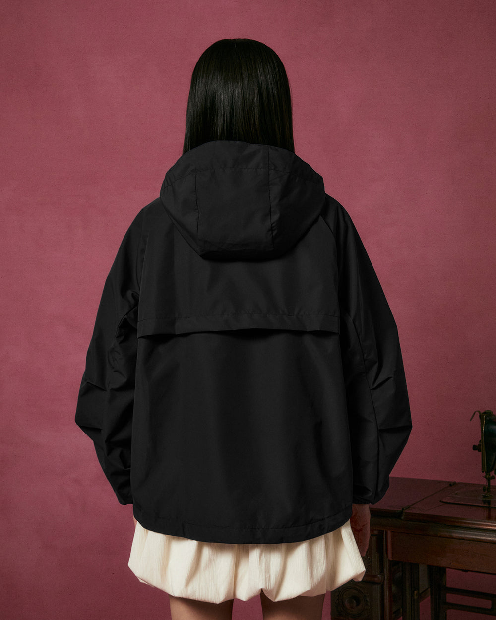 A person with long dark hair, dressed in the Sundae School BLACK JEOGORI TIE SHELL JACKET and a white skirt, stands facing away against a pink background beside a wooden table.