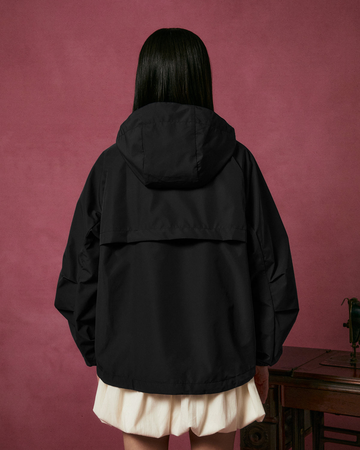 A person with long dark hair, dressed in the Sundae School BLACK JEOGORI TIE SHELL JACKET and a white skirt, stands facing away against a pink background beside a wooden table.
