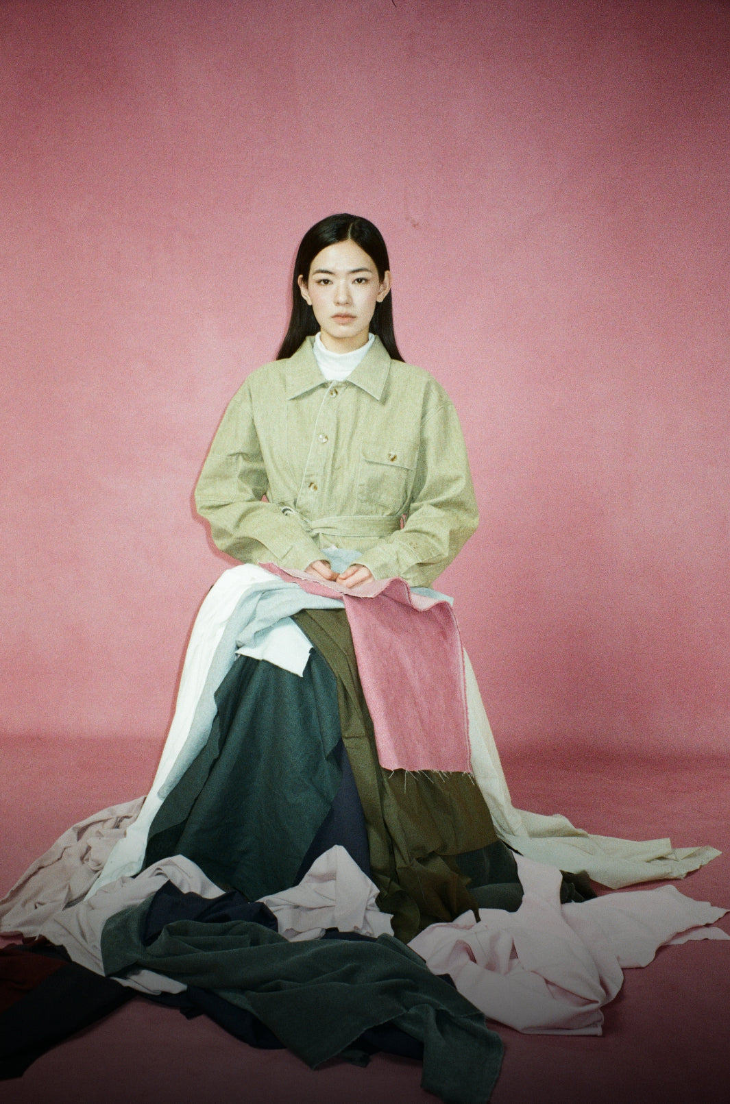 A model sits against a textured pink studio backdrop wearing a light sage green jacket over a white mock neck top, layered with a long, wide-leg skirt in multiple colors including pink, dark green, and cream. Scattered fabric pieces are on the floor around her.