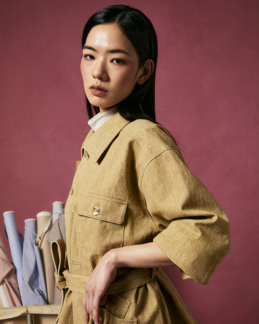 A woman with straight black hair wears the Sundae School TAN ASYMMETRIC WORK JACKET, belted at the waist, standing against a dark pink background. Pastel rolled fabrics are stacked behind her on the left as she looks at the camera.