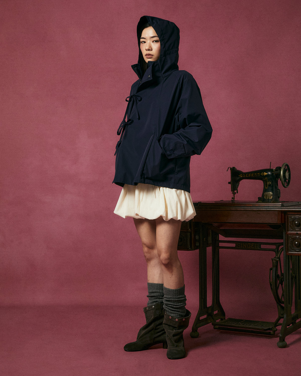 A person wearing the Sundae School NAVY JEOGORI TIE SHELL JACKET, a cream skirt, gray socks, and brown boots stands next to an antique sewing machine against a pink background.