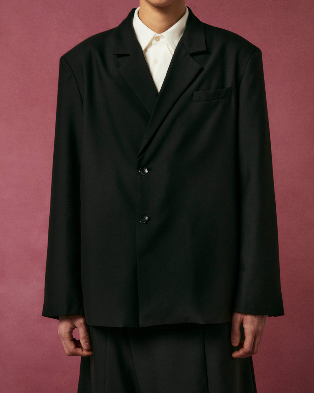 A person wears the BLACK DOUBLE DUTY BLAZER by Sundae School over a white button-up shirt, standing against a muted pink background with their face partially out of frame.