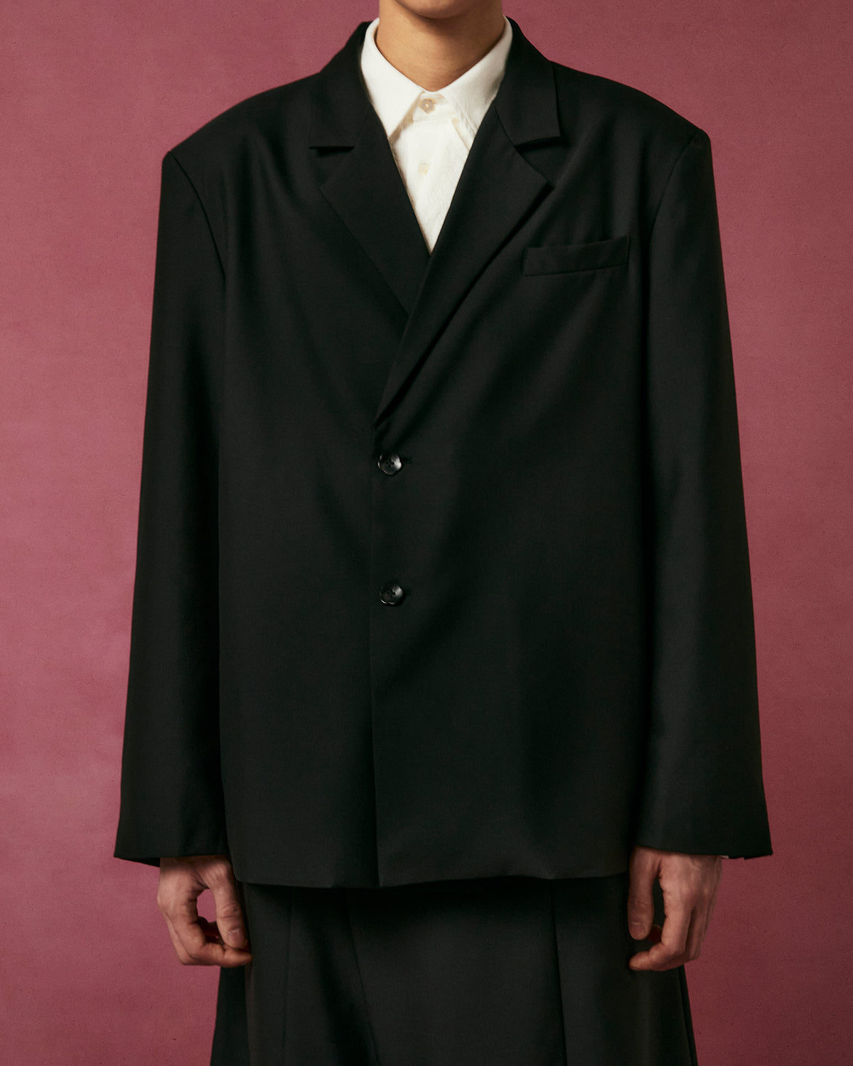 A person wears the BLACK DOUBLE DUTY BLAZER by Sundae School over a white button-up shirt, standing against a muted pink background with their face partially out of frame.