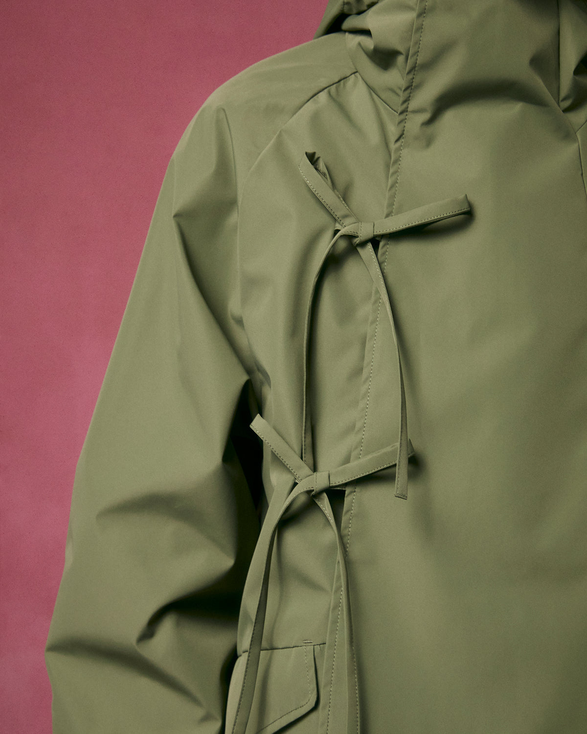 Close-up of the Sundae School MATCHA JEOGORI TIE SHELL JACKET in olive green, featuring two front fabric ties and a visible sleeve, set against a muted pink background. Inspired by traditional Korean design.