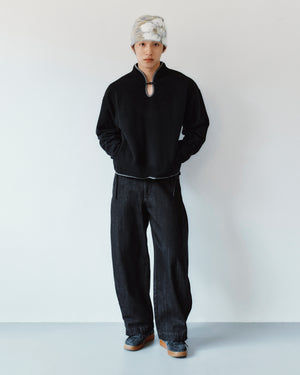 A person wearing a black mock-neck sweatshirt with a keyhole detail and BLACK ACCORDION DENIM PANTS in black.