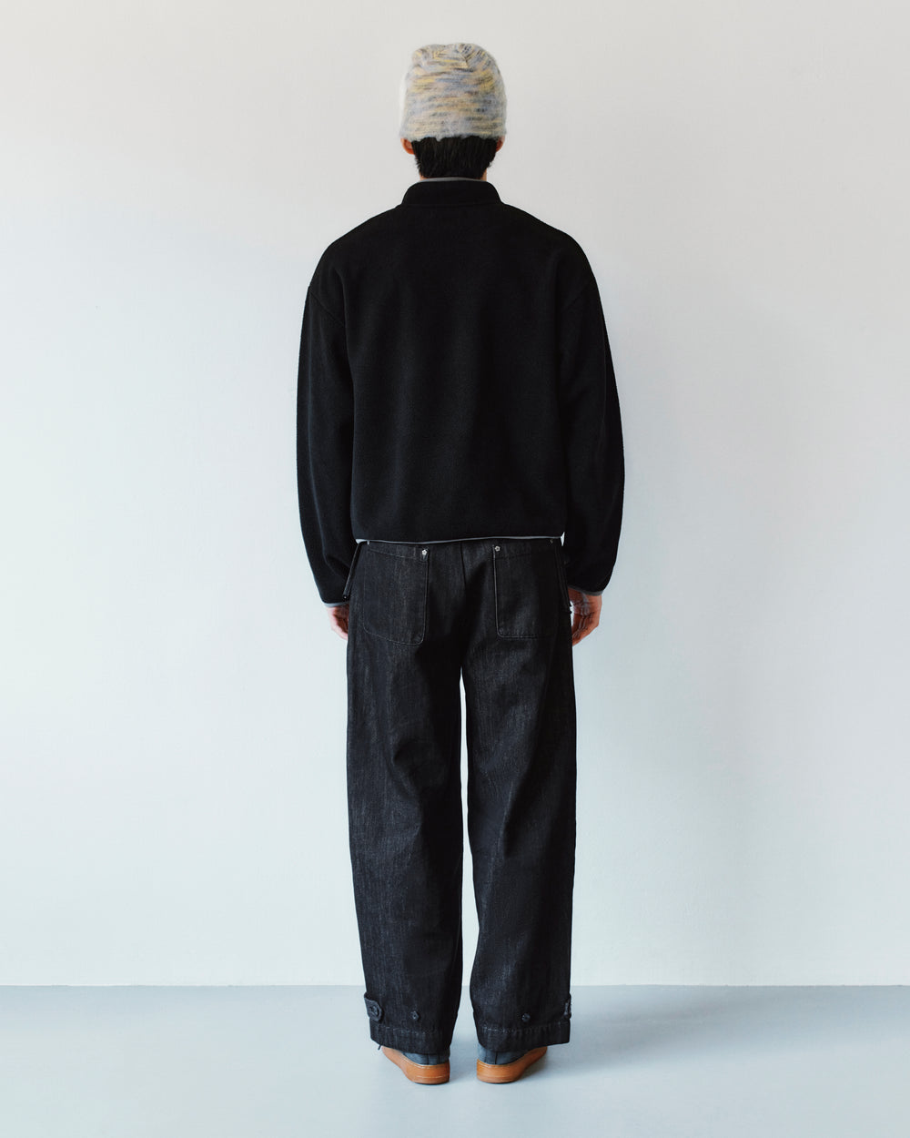 A person seen from behind, wearing a black jacket and Sundae School BLACK ACCORDION DENIM PANTS, which are loose-fitting black denim flares with buttoned cuffs.