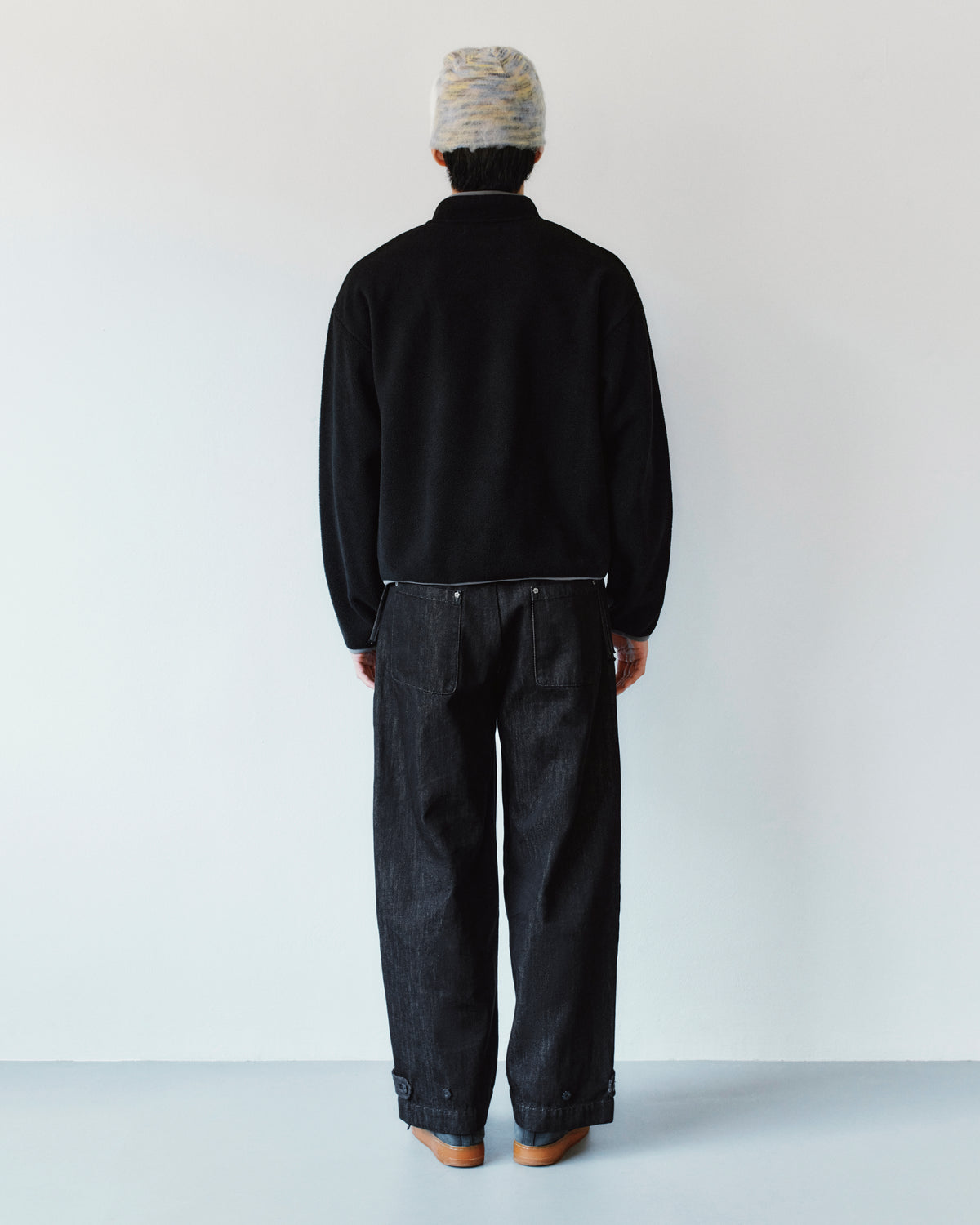 A person seen from behind, wearing a black jacket and Sundae School BLACK ACCORDION DENIM PANTS, which are loose-fitting black denim flares with buttoned cuffs.