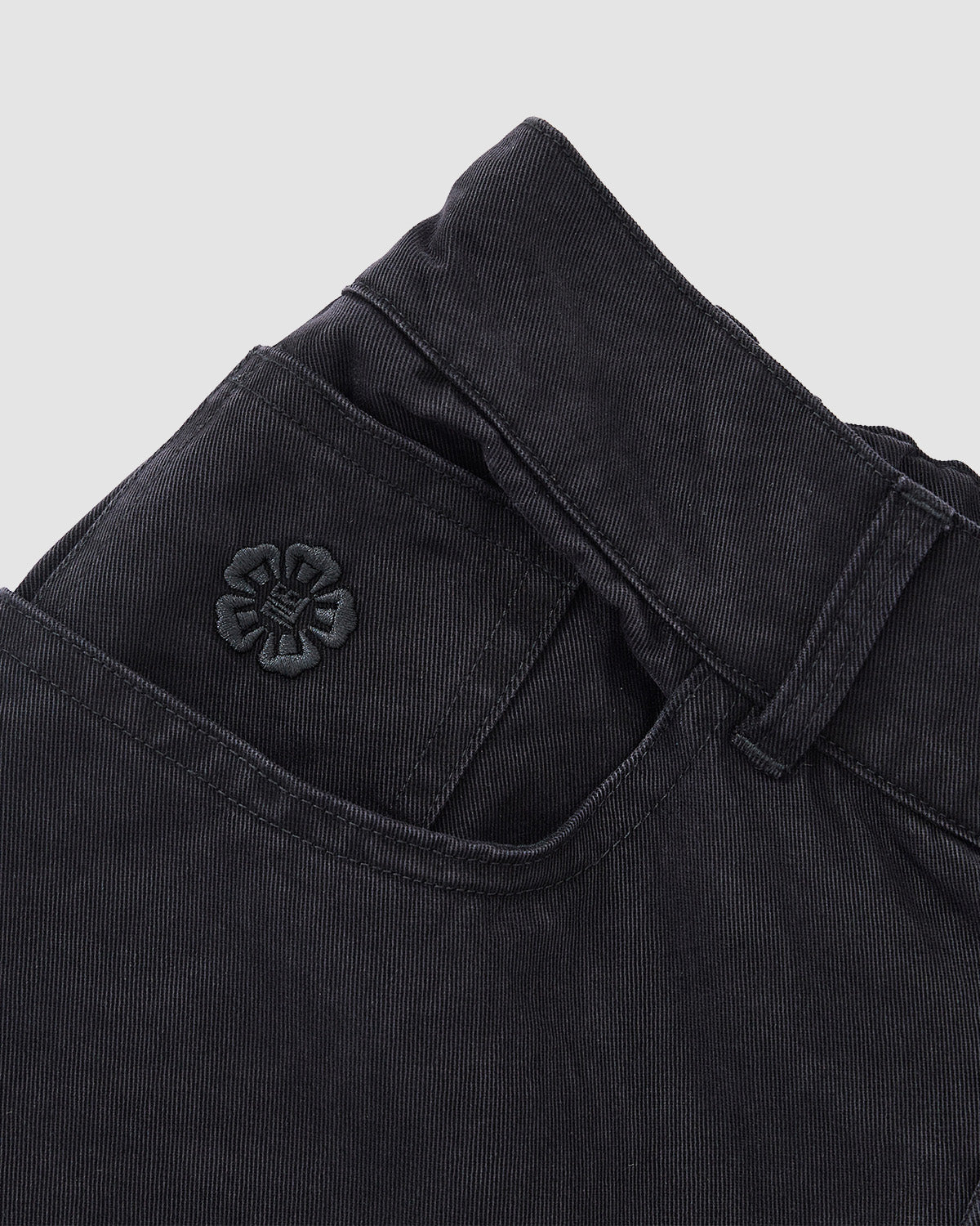 Close-up of the waistband and pocket of Sundae School BLACK ACCORDION CHINO PANTS, featuring a black embroidered flower above the pocket and hidden accordion pleats, shown on a light gray background.