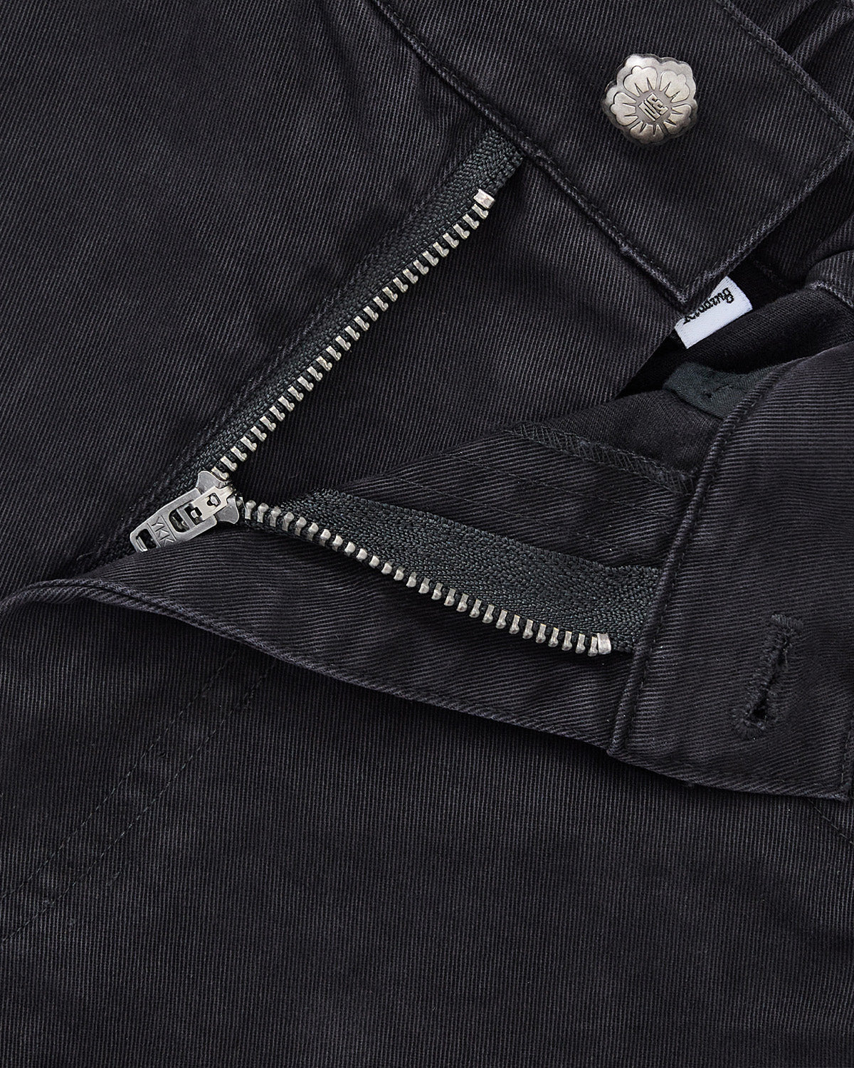 Close-up of Sundae School’s BLACK ACCORDION CHINO PANTS in black corduroy, showing pleats, a partially unzipped metal zipper, silver flower-shaped button, and a small inner brand tag.