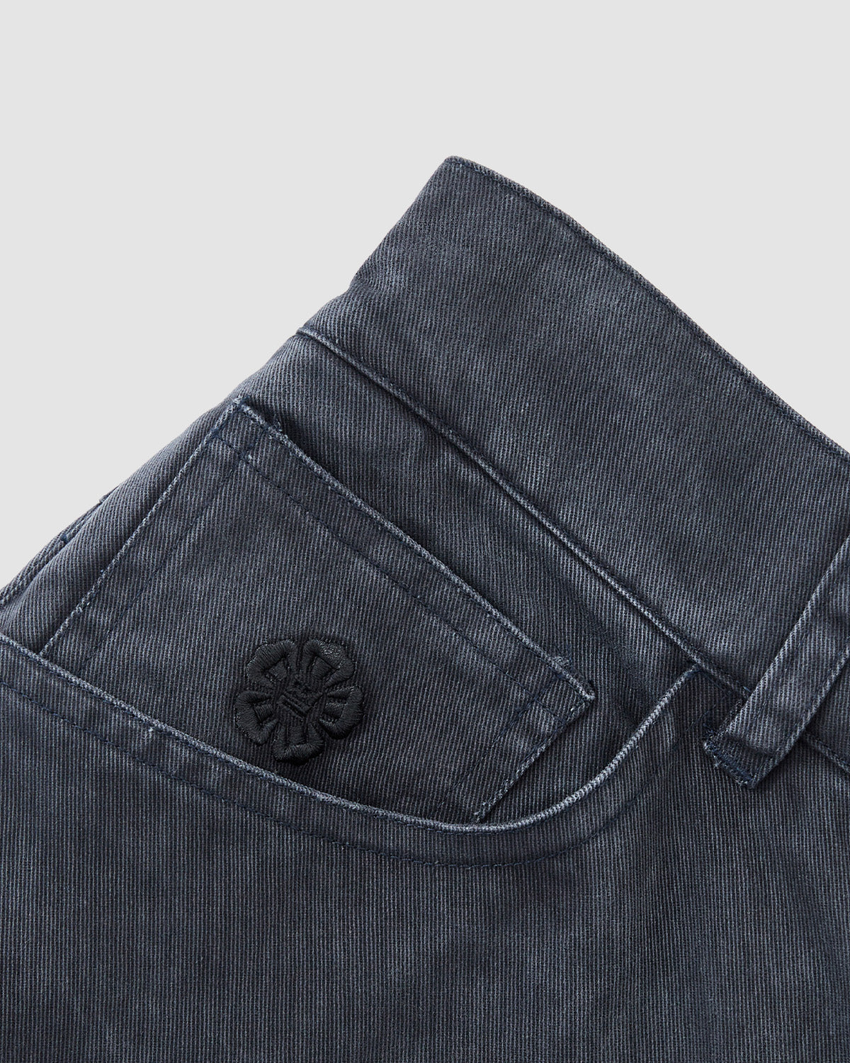 Close-up of Sundae School CHARCOAL ACCORDION CHINO PANTS pocket with flap, belt loop, and an embroidered circular black patch with a geometric pattern, inspired by hanbok. Light gray background.