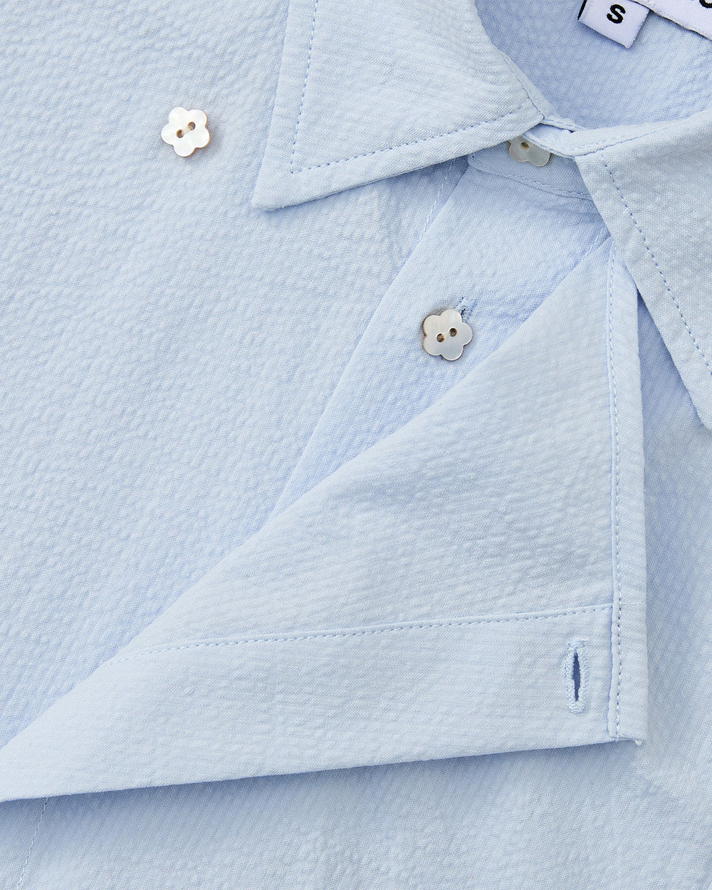 A close-up of the Sundae School SKY BLUE TIE GORUM SHIRT shows its folded collar, off-white flower-shaped buttons, and a visible 'S' tag indicating size. The shirt features light blue seersucker cotton fabric.